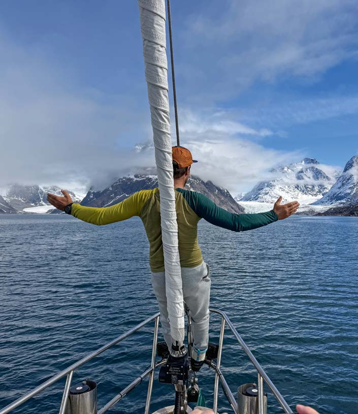 Mann steht mit ausgebreiteten Armen am Bug eines Segelschiffs im Fjord