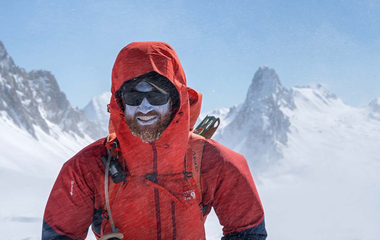 Porträt eines bärtigen Bergsteigers in roter Kapuzenjacke vor spitzen Granitgipfeln