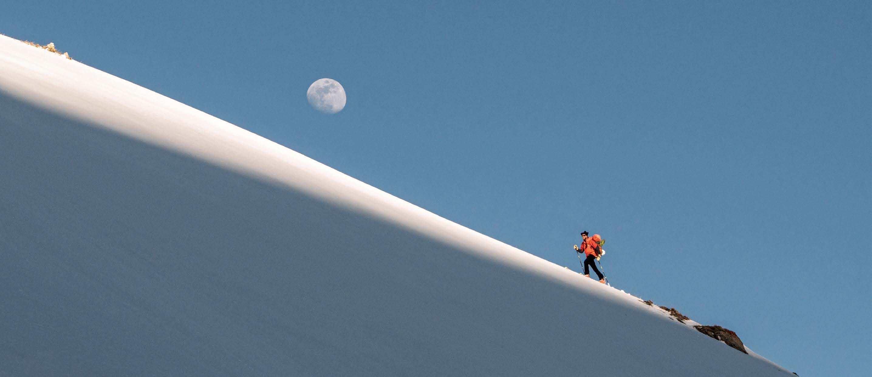 Skitourengeher steigt einen steilen Schneegrat unter blauem Himmel mit Mond auf