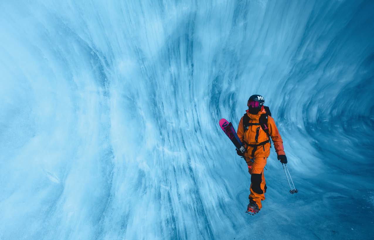 Skifahrer in oranger Jacke steht mit Eisgerät in einem leuchtend blauen Eistunnel