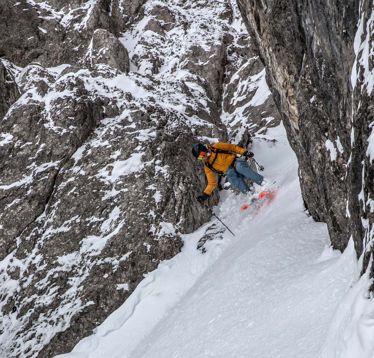 Skifahrerin fährt in gelber Jacke neben einer Felswand durch das steile Couloir ab