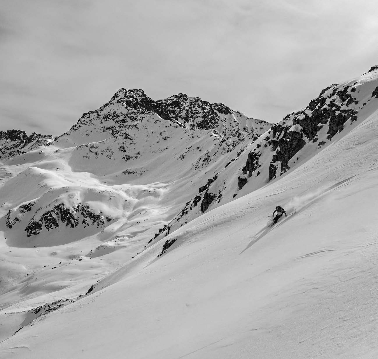 Schwarz-Weiß-Aufnahme eines Skifahrers in einem Pulverhang unter Felsgipfel