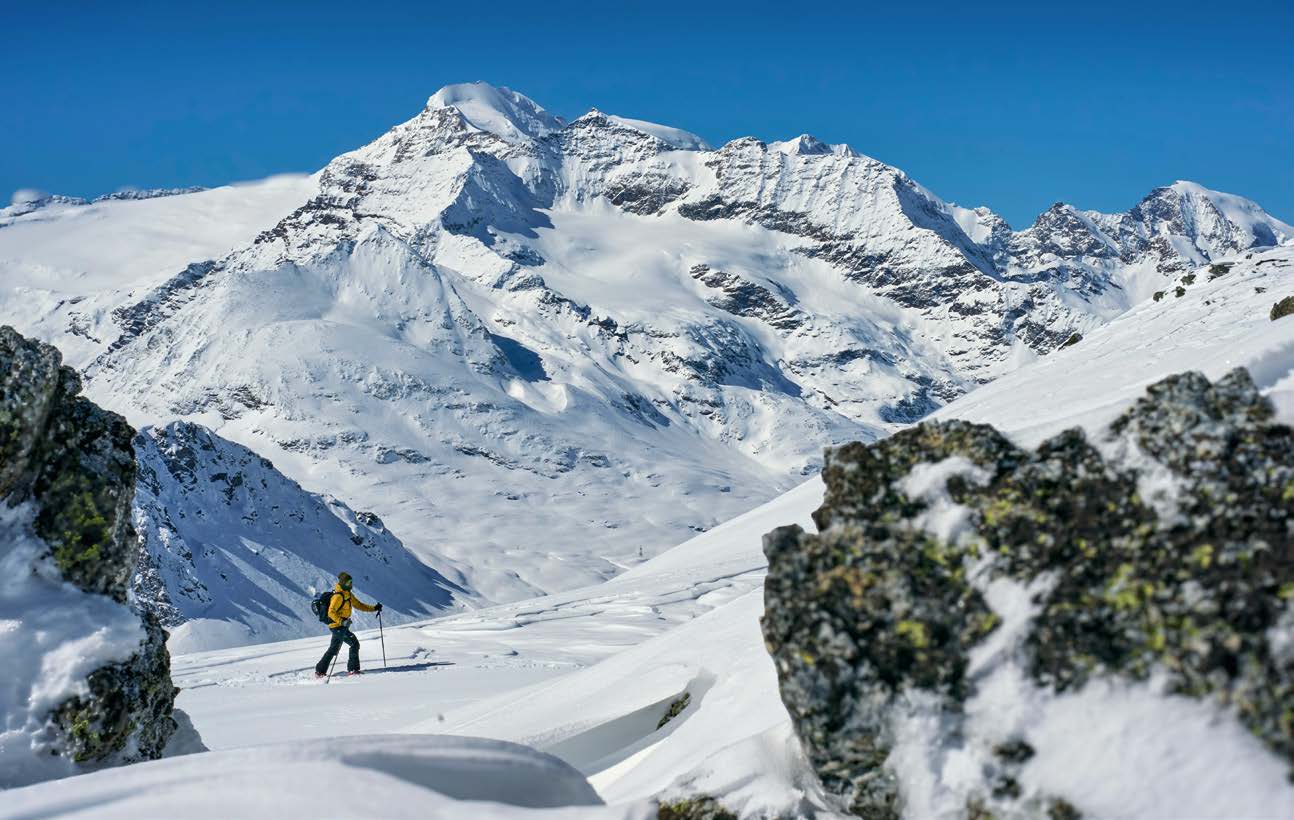 Skitourengeher steigt zwischen flechtenbewachsenen Felsen vor vergletschertem Gipfel auf