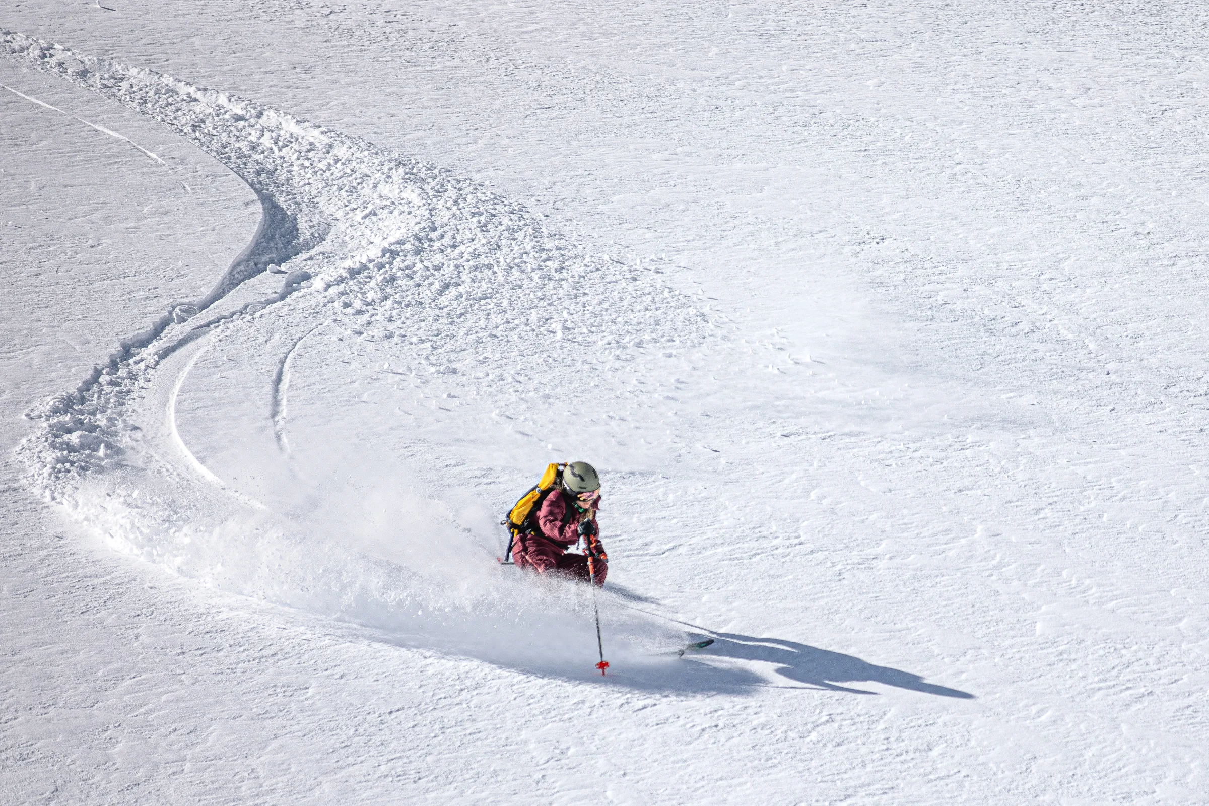 Andrea Limbacher turns through deep snow on an open slope in Gastein, wearing a yellow backpack and leaving a curved track behind her.