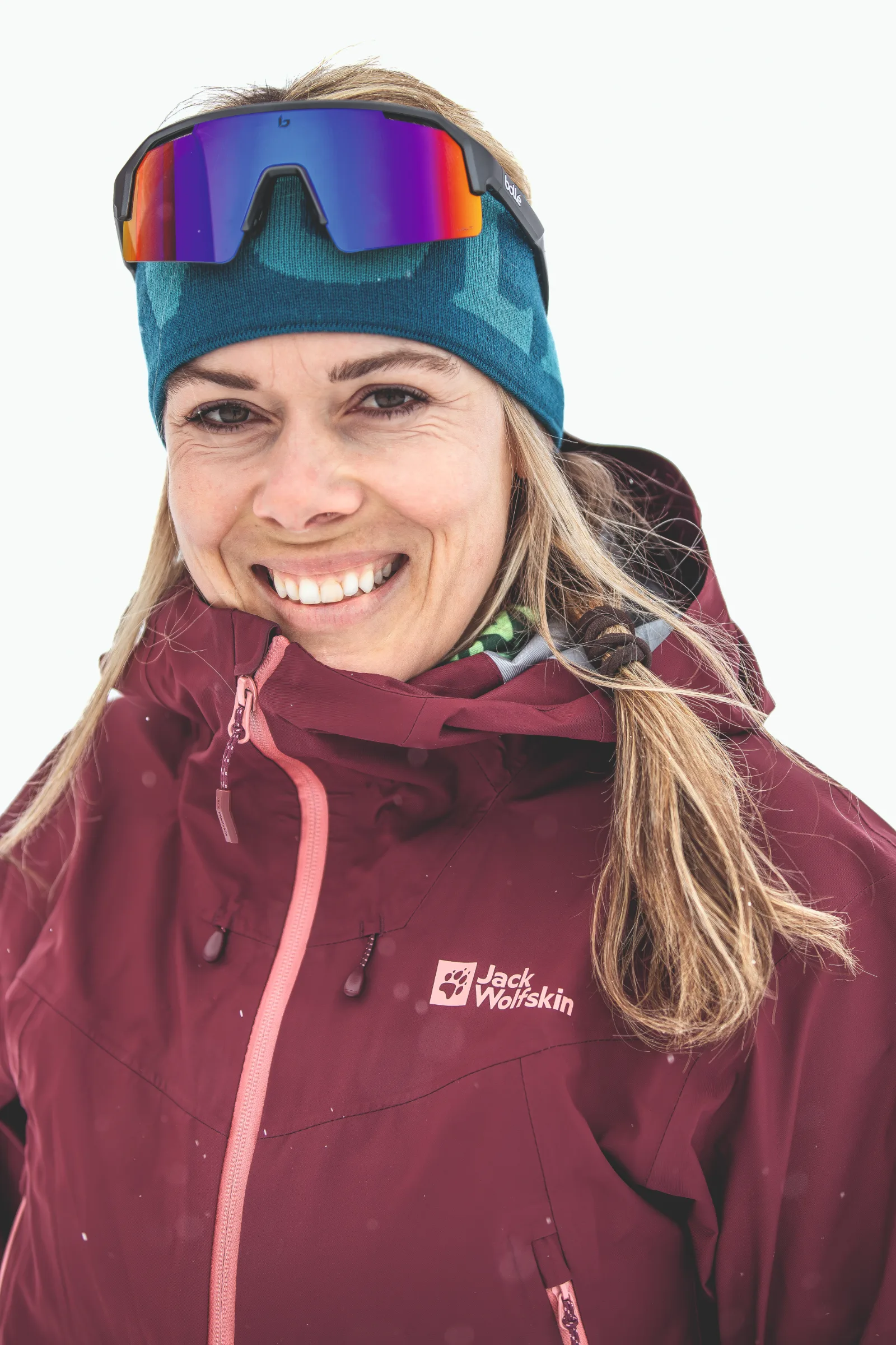Andrea Limbacher smiles in a close-up against a white background, wearing a teal headband with mirrored sports sunglasses resting on it and a burgundy jacket.