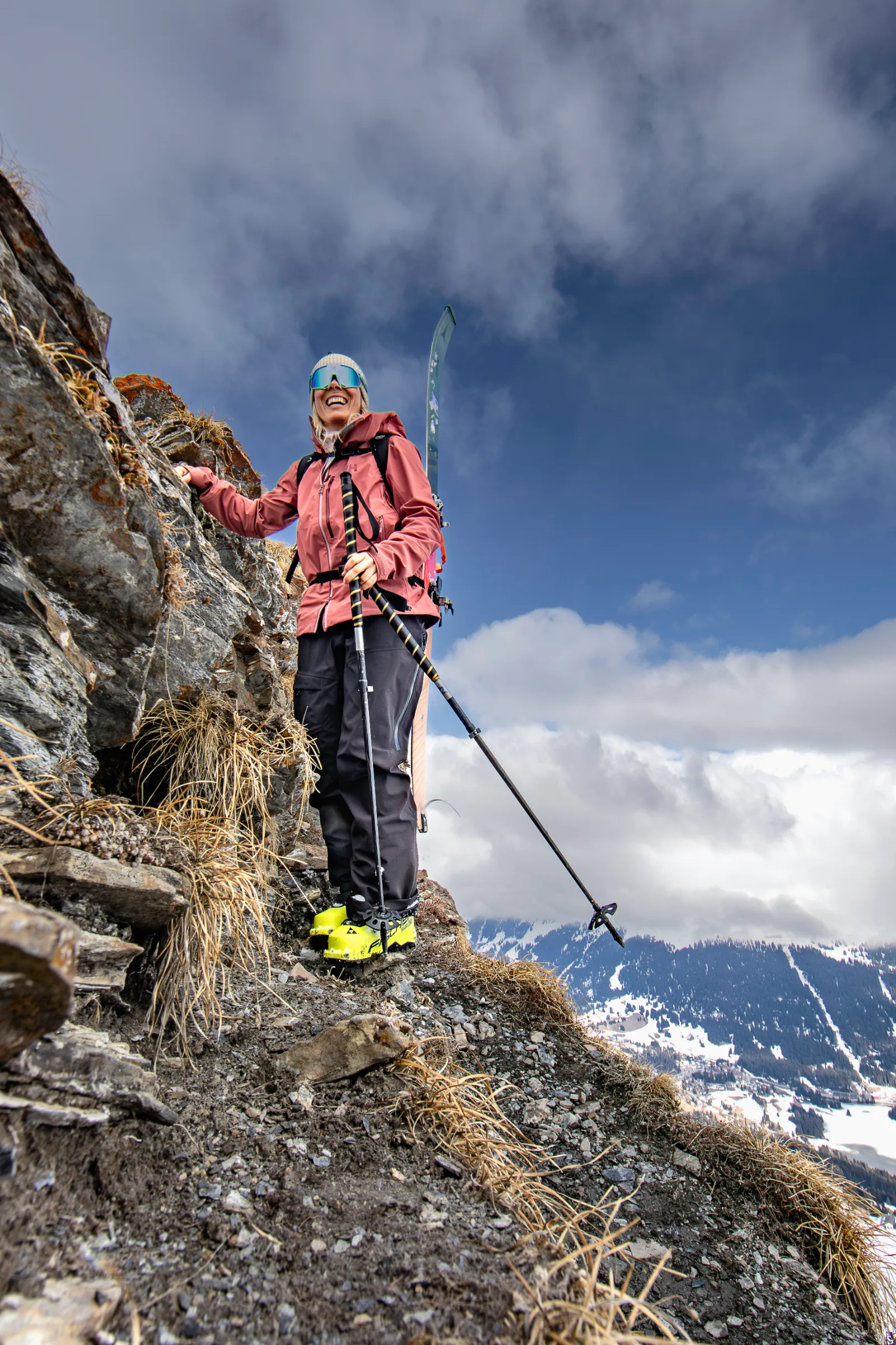 Andrea Limbacher standing in ski boots with ski poles on a narrow rocky path above snow-covered mountains in Lenzerheide.