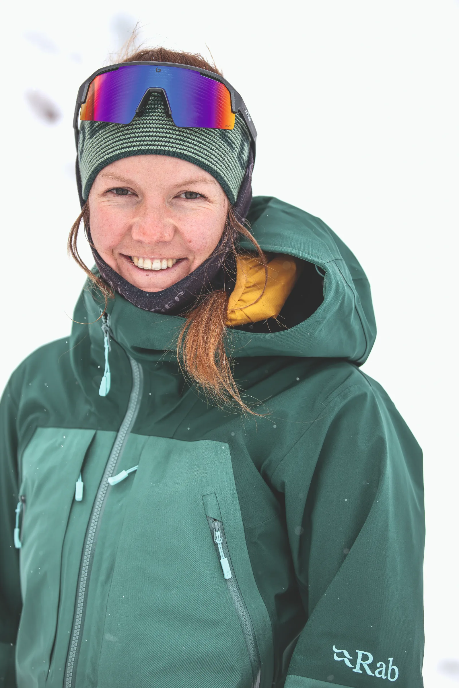 Annika Schlachter smiles at the camera against a snowy background, wearing a green jacket, a striped headband and mirrored sports sunglasses pushed onto her forehead.