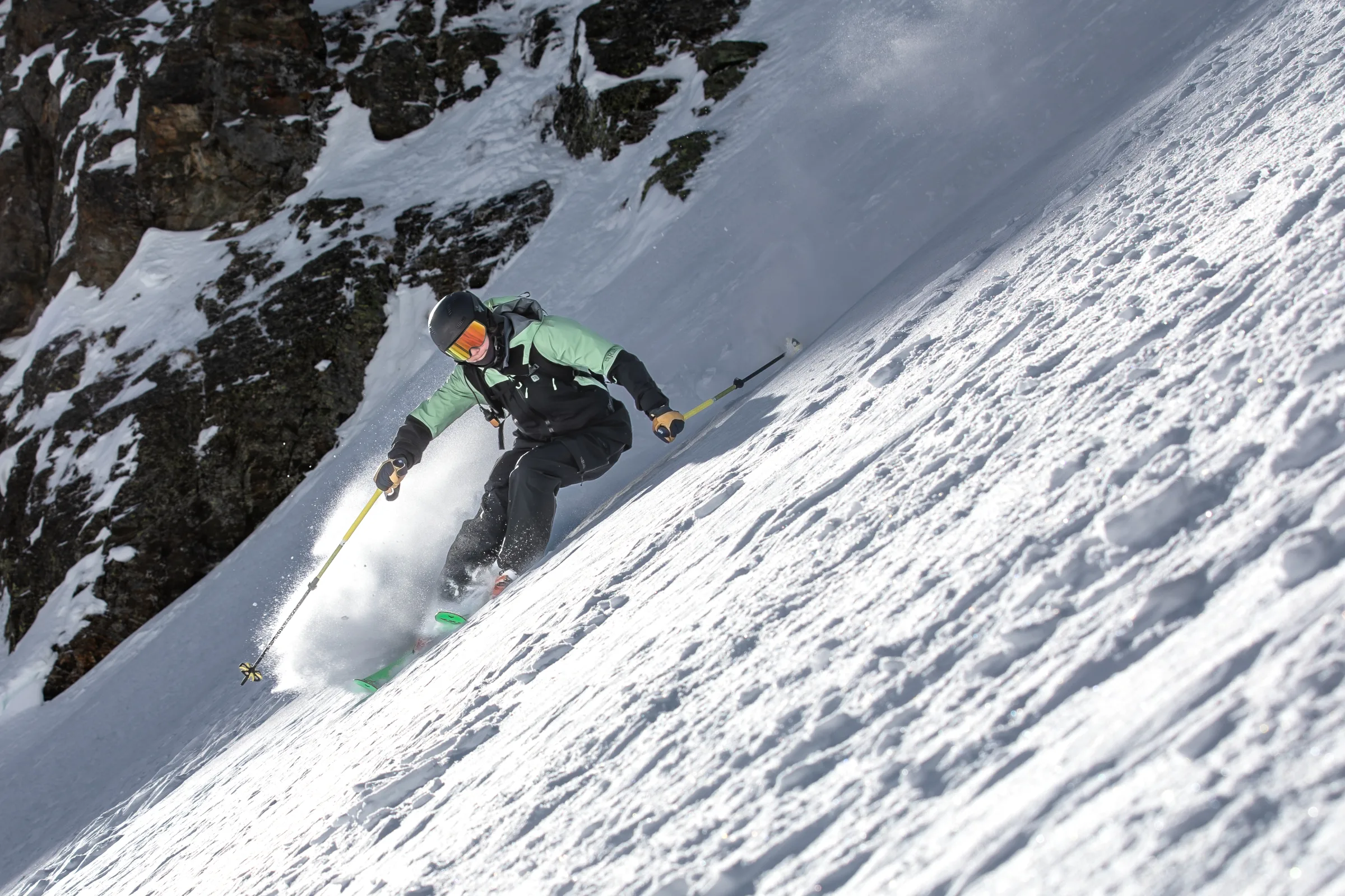 Annika Schlachter descends a steep snowy slope beside dark rock walls on green skis, holding yellow poles and kicking up snow.