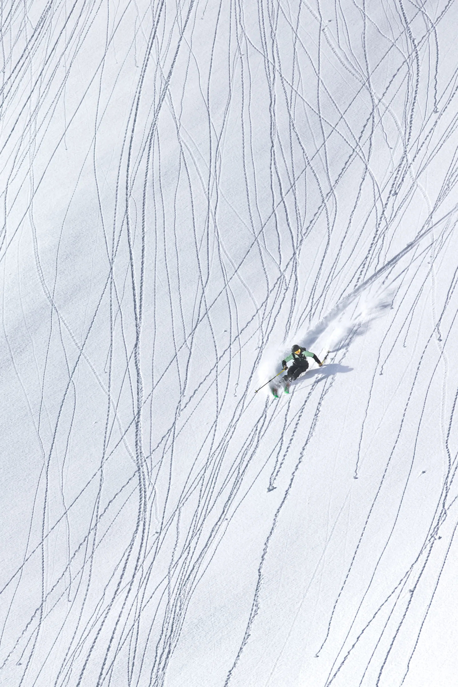 Viewed from above, Annika Schlachter skis down a slope crisscrossed by narrow tracks, trailing a plume of snow behind her green skis.
