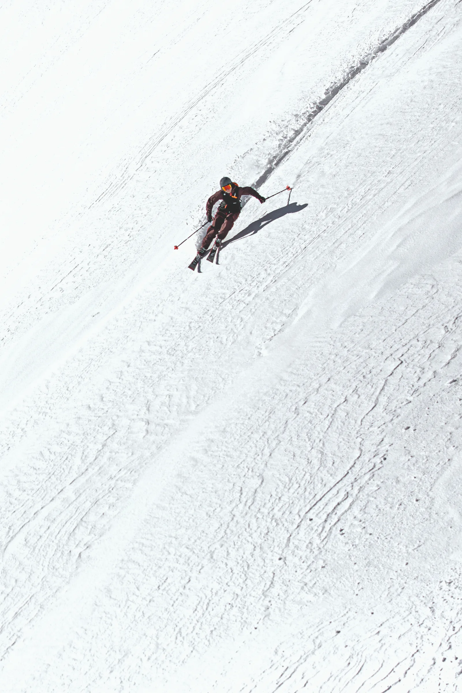 Bernd Hassmann skis down a broad snowy slope with his poles held out to the sides, leaving a track behind him and casting a distinct shadow.