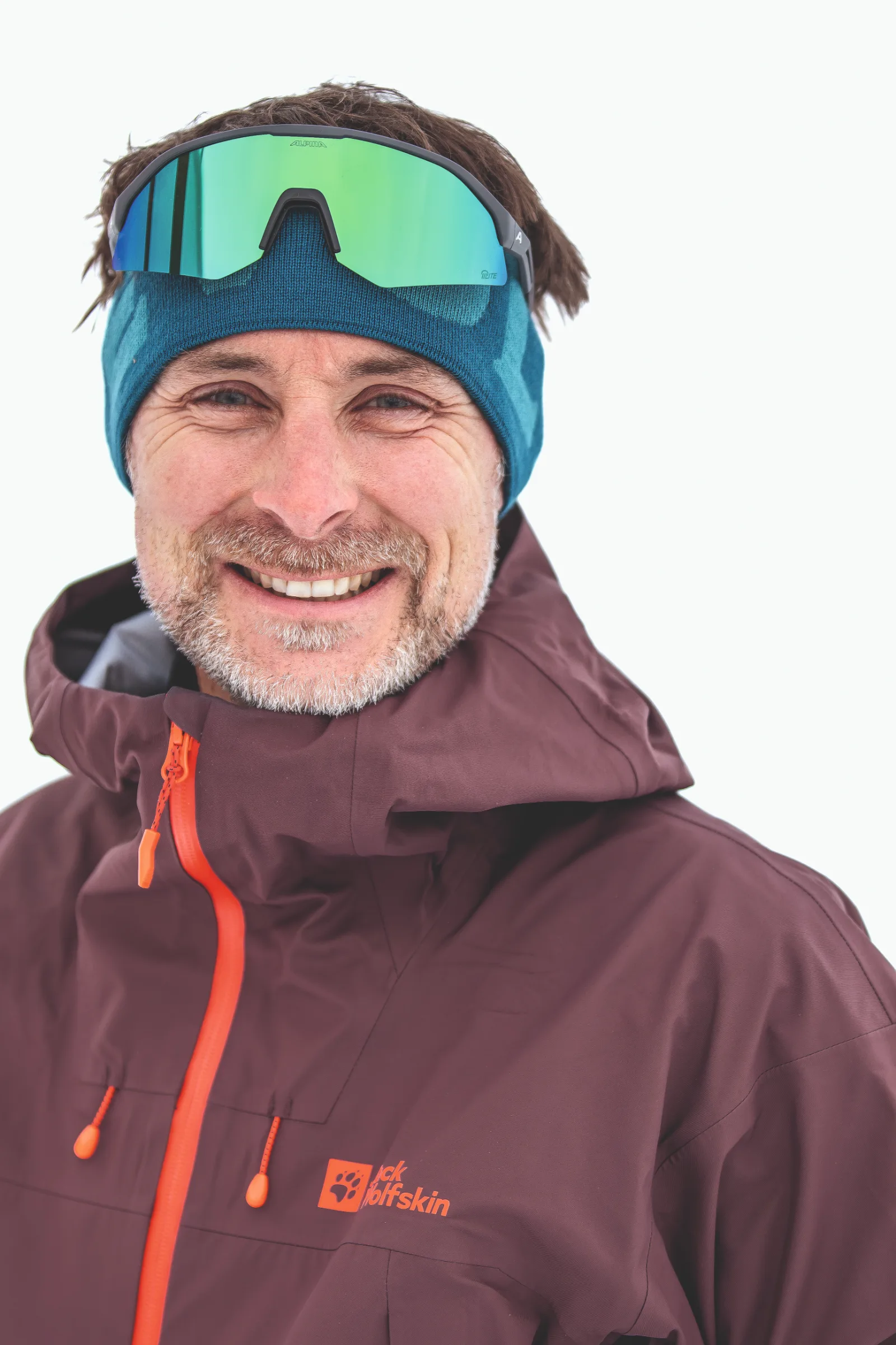 Bernd Hassmann smiles at the camera against a white background, wearing a blue headband, green mirrored sports glasses pushed up on his forehead and a burgundy jacket.