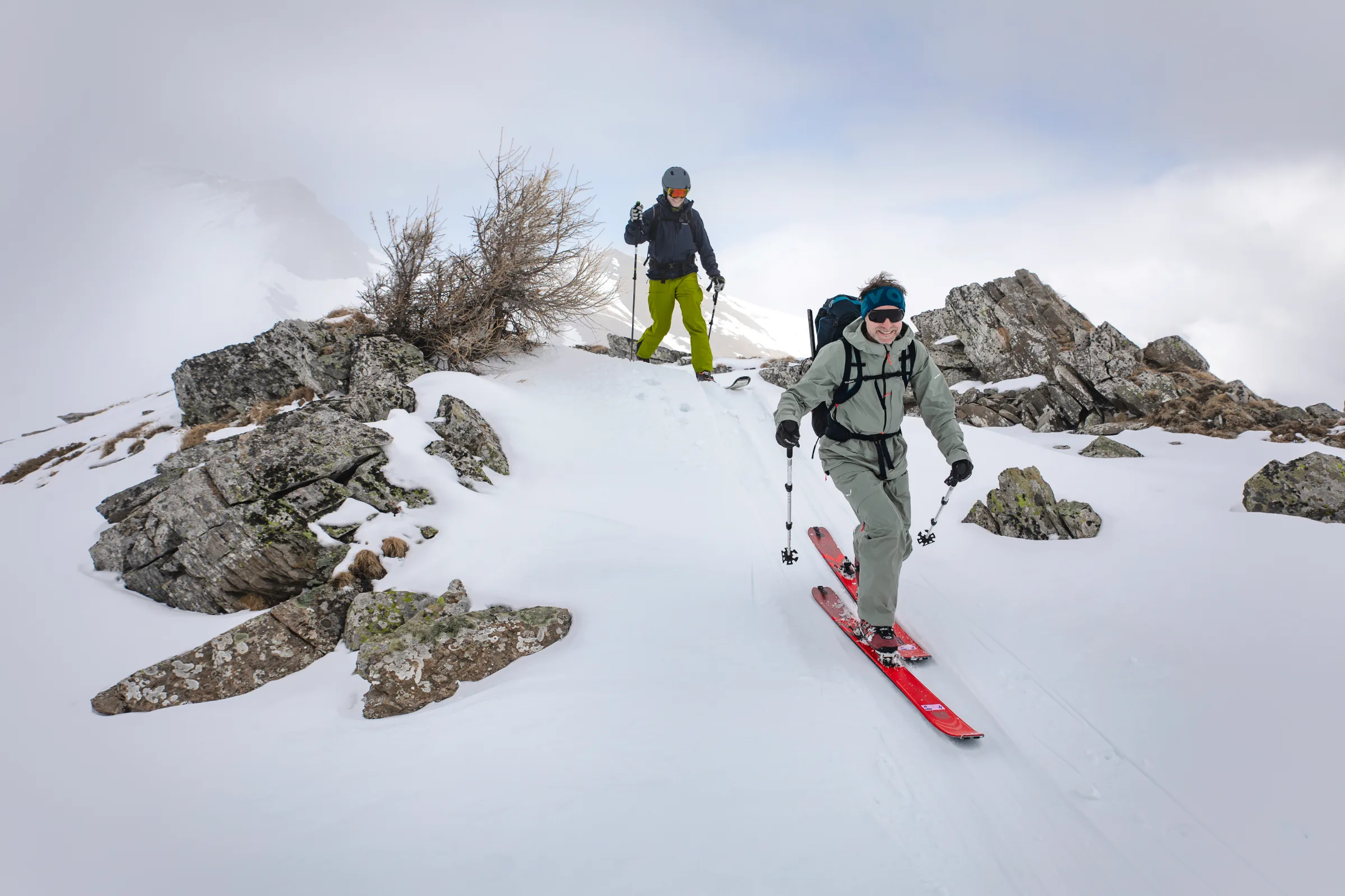 Bernd Hassmann moves on red skis between rocks along a snowy ridge near Lenzerheide, followed by another person in the mist.
