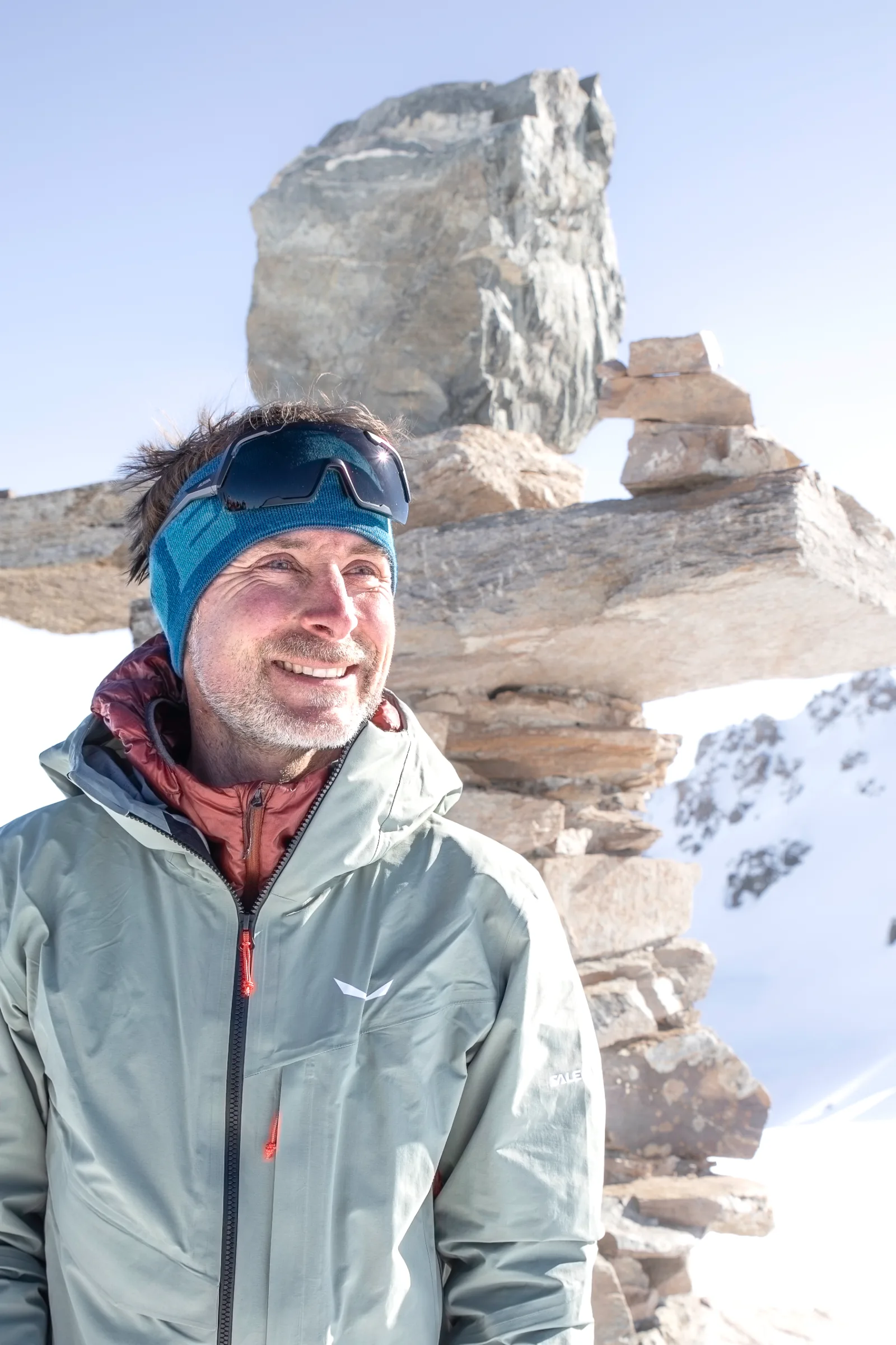 Bernd Hassmann smiles with a blue headband and sunglasses pushed up on his forehead, with stacked stone slabs and snowy mountains behind him.