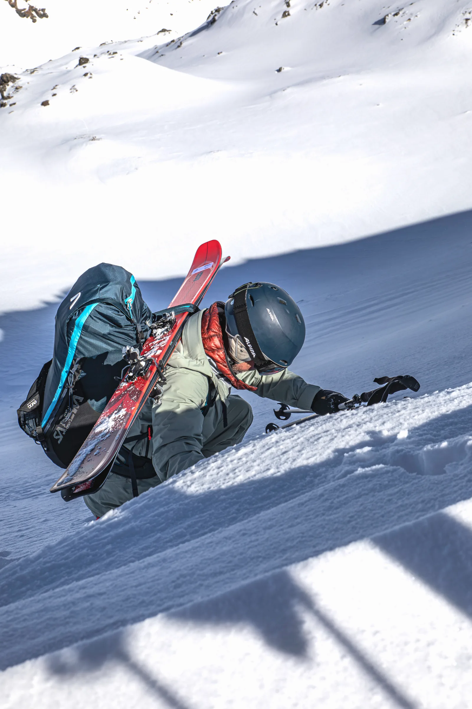 Bernd Hassmann climbs a steep snowy slope with a helmet and red skis strapped to his backpack, bracing one hand in the snow.