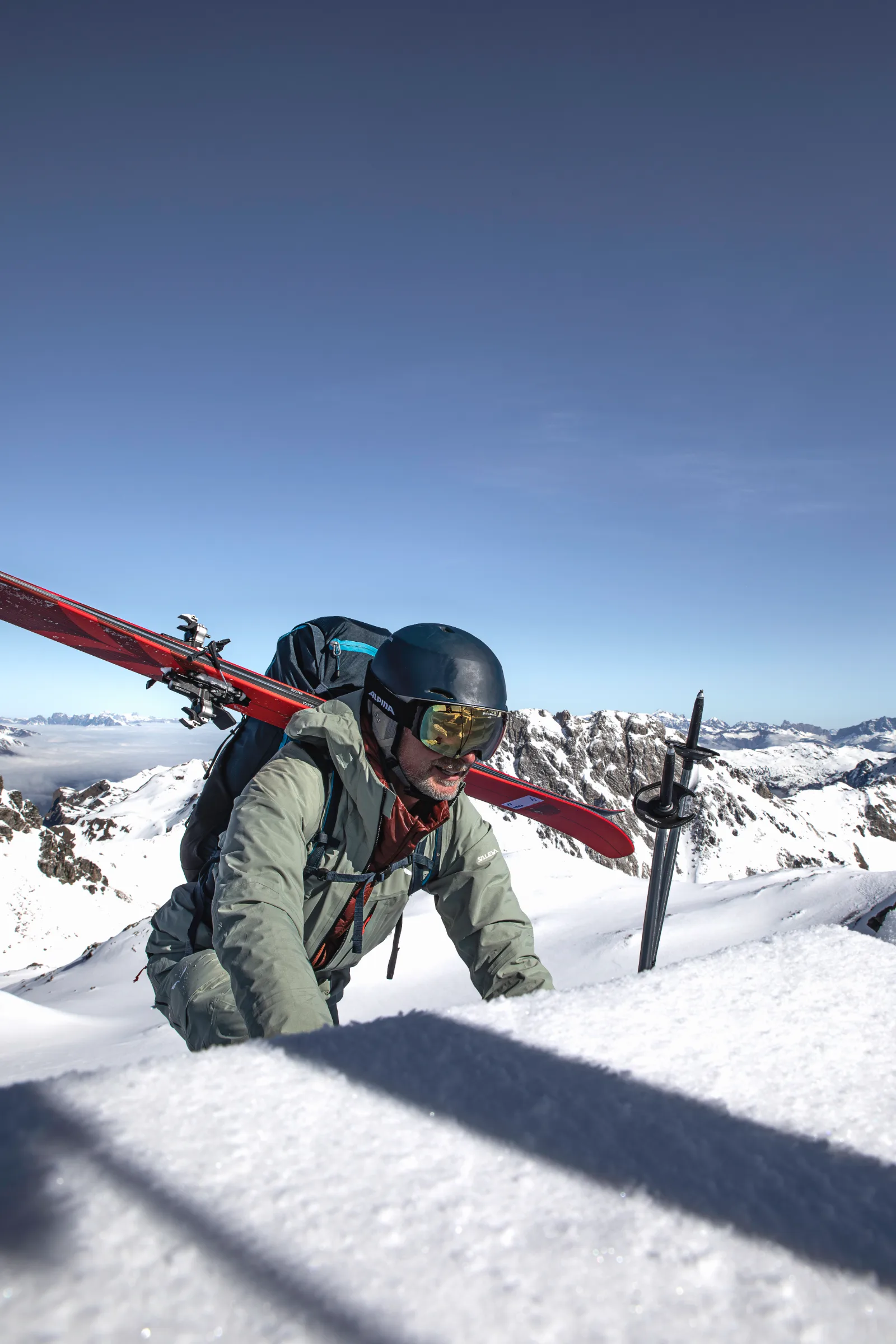 Bernd Hassmann climbs over a snowy lip with red skis across his backpack; poles stand beside him and snowy peaks rise beneath a blue sky.