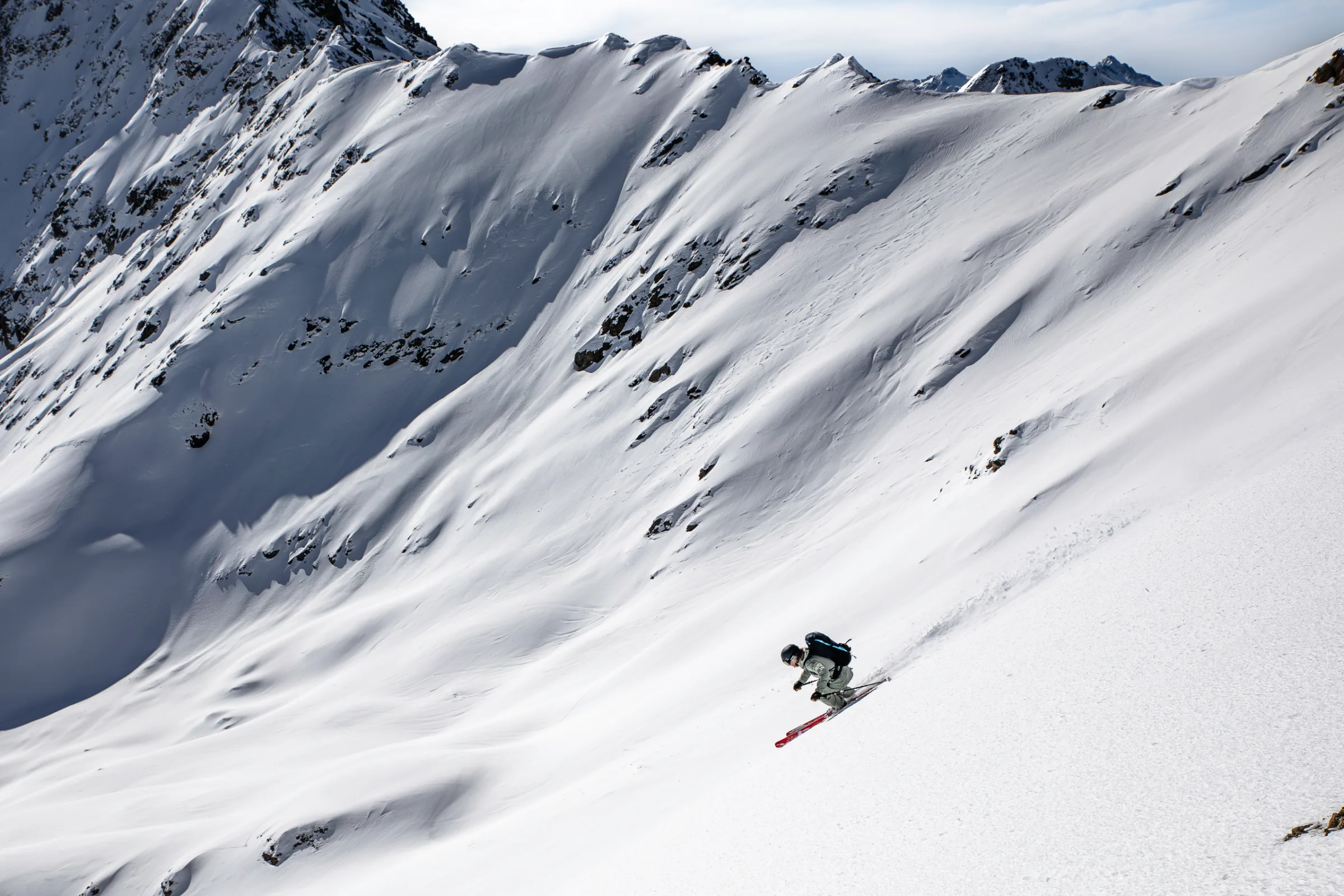 Bernd Hassmann descends a steep slope on red skis in a low crouch, against a broad mountainside covered in snow and exposed rocks.