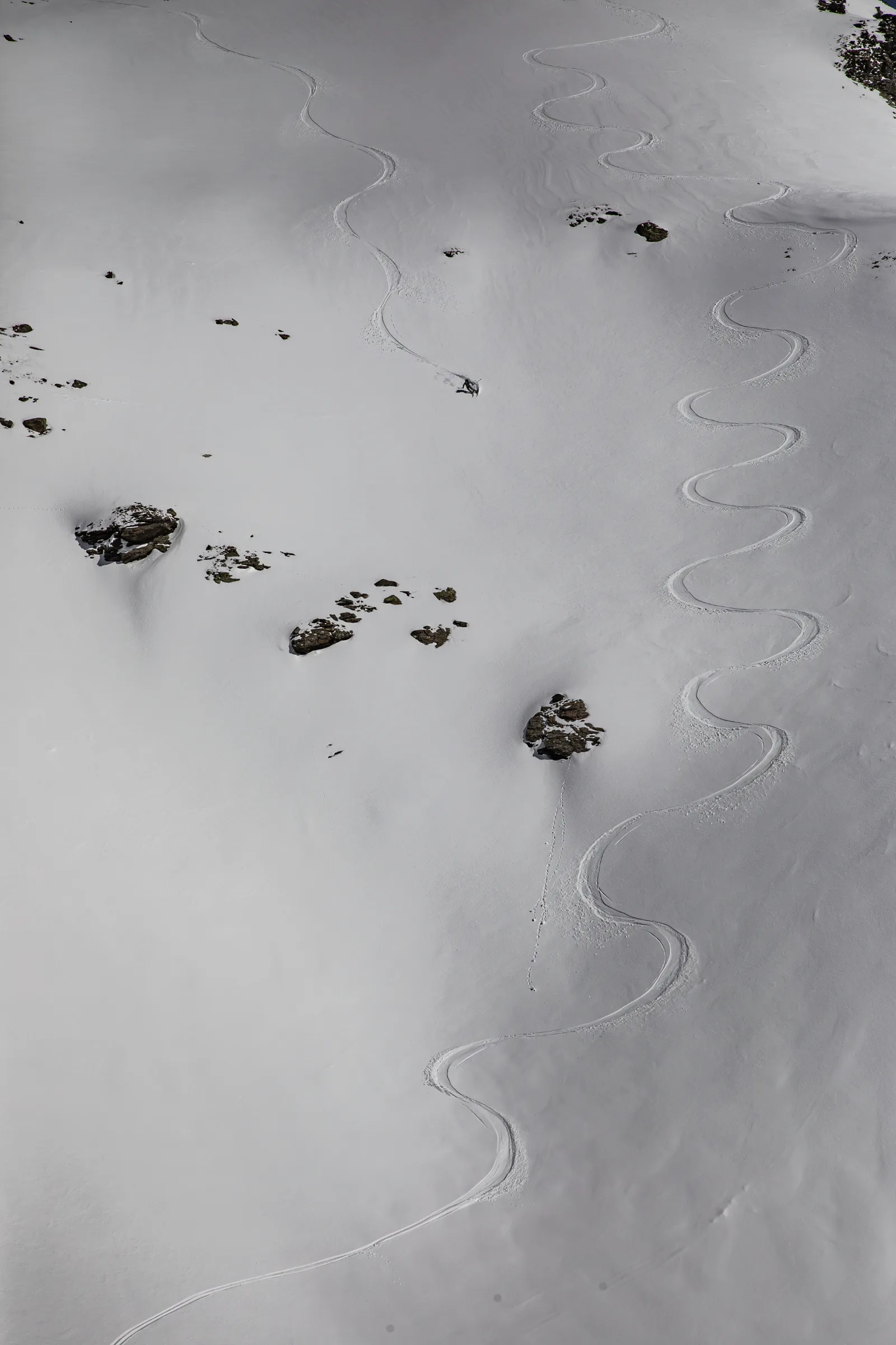 A lone skier makes turns down a broad, rock-studded snow slope beside a long winding ski track.