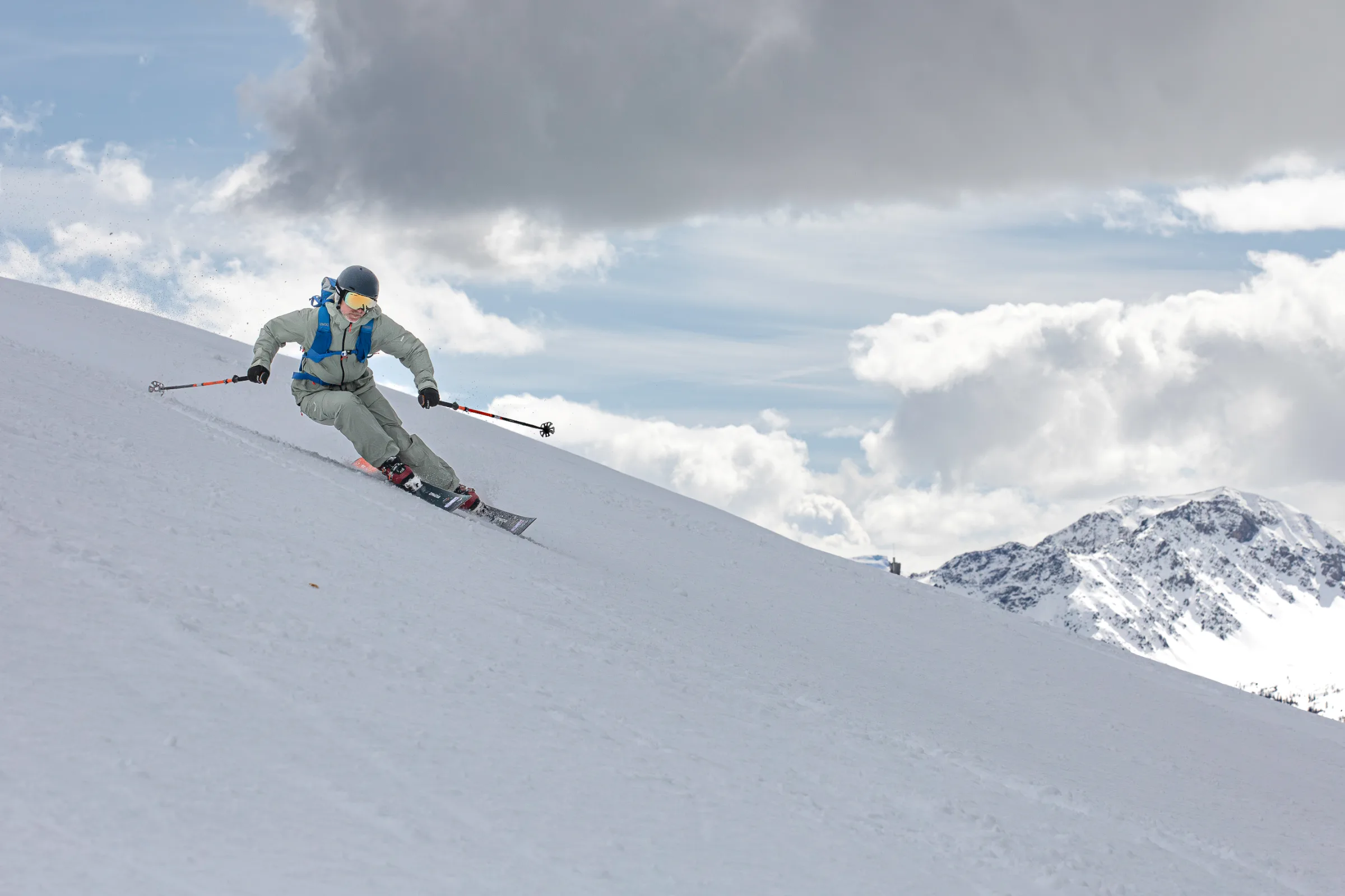 Bernd Hassmann skis down a snowy slope in Lenzerheide wearing light grey clothing and a blue backpack, with clouds and snow-covered mountains behind him.