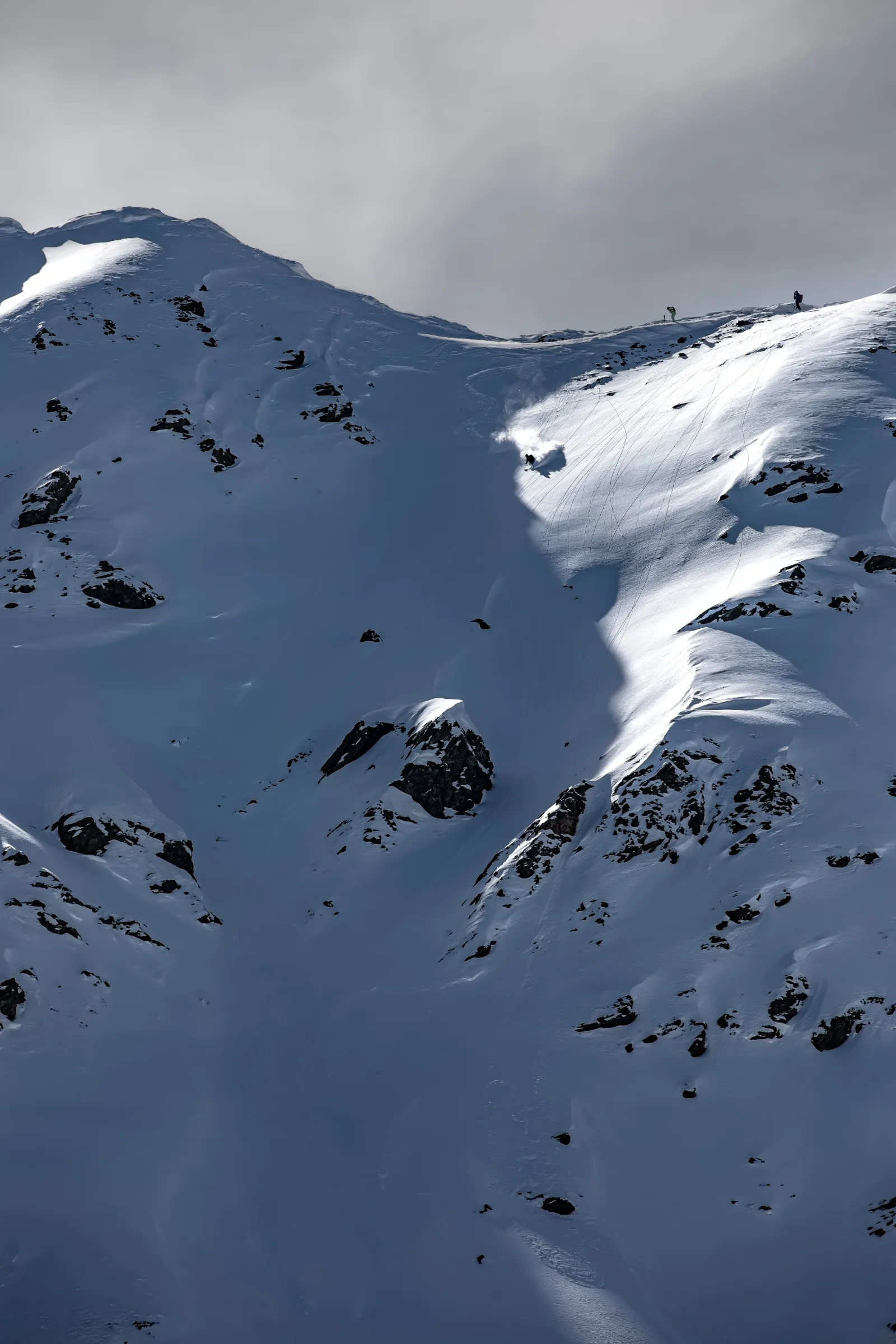 A skier throws up snow while turning down a steep, rocky slope, with two people standing on the ridge above.
