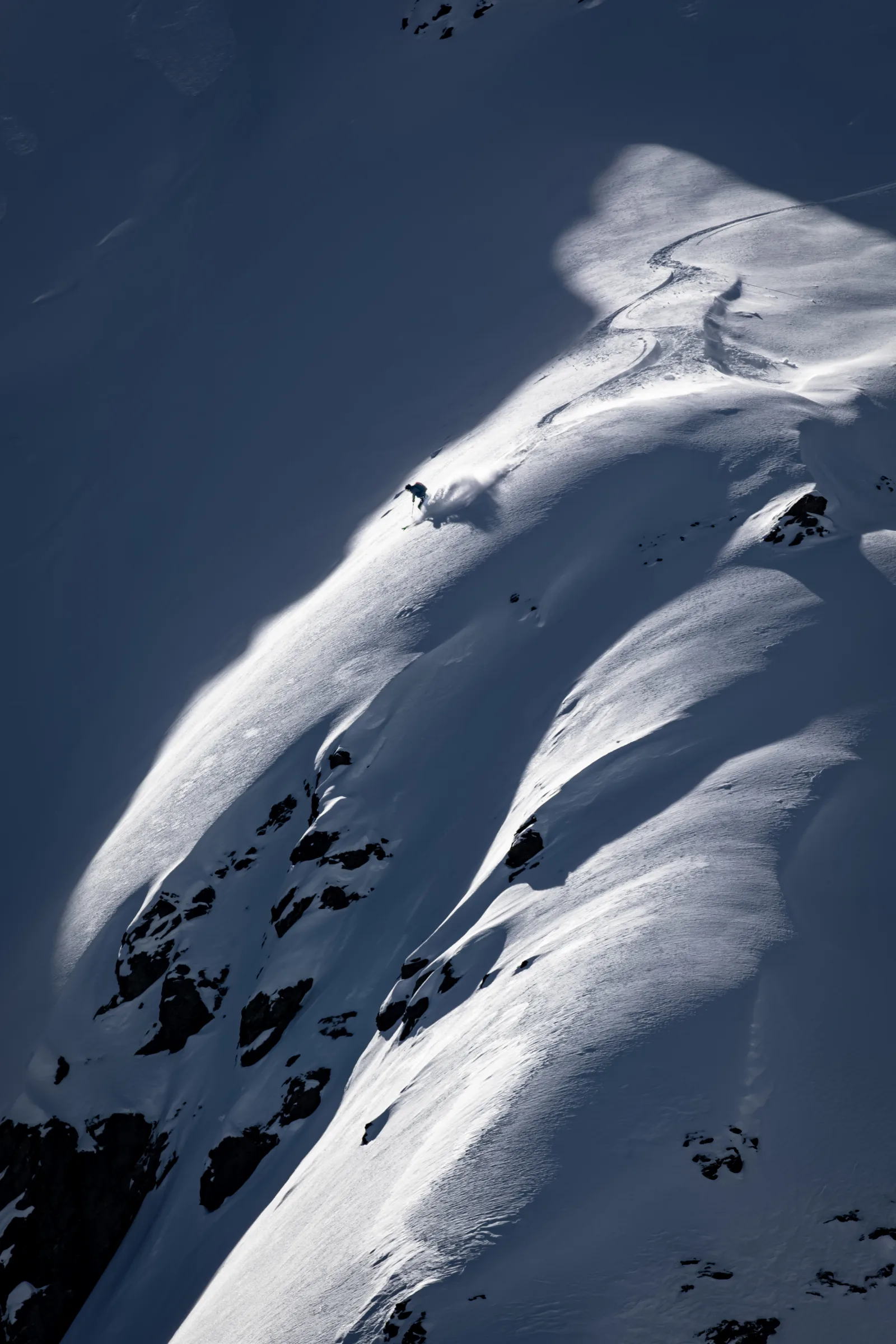 A skier descends a narrow sunlit strip on a steep snowy slope, leaving curved tracks between deep shadows and exposed rocks.