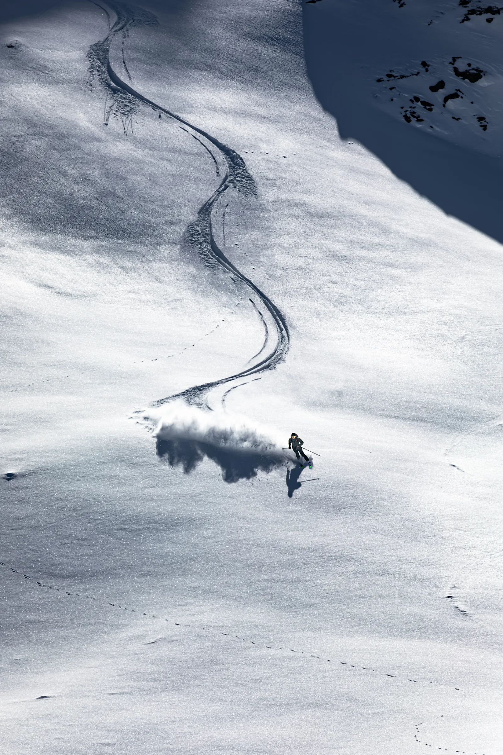 A skier throws up a broad plume of snow on an open, sunlit slope, with a long winding track stretching uphill behind him.