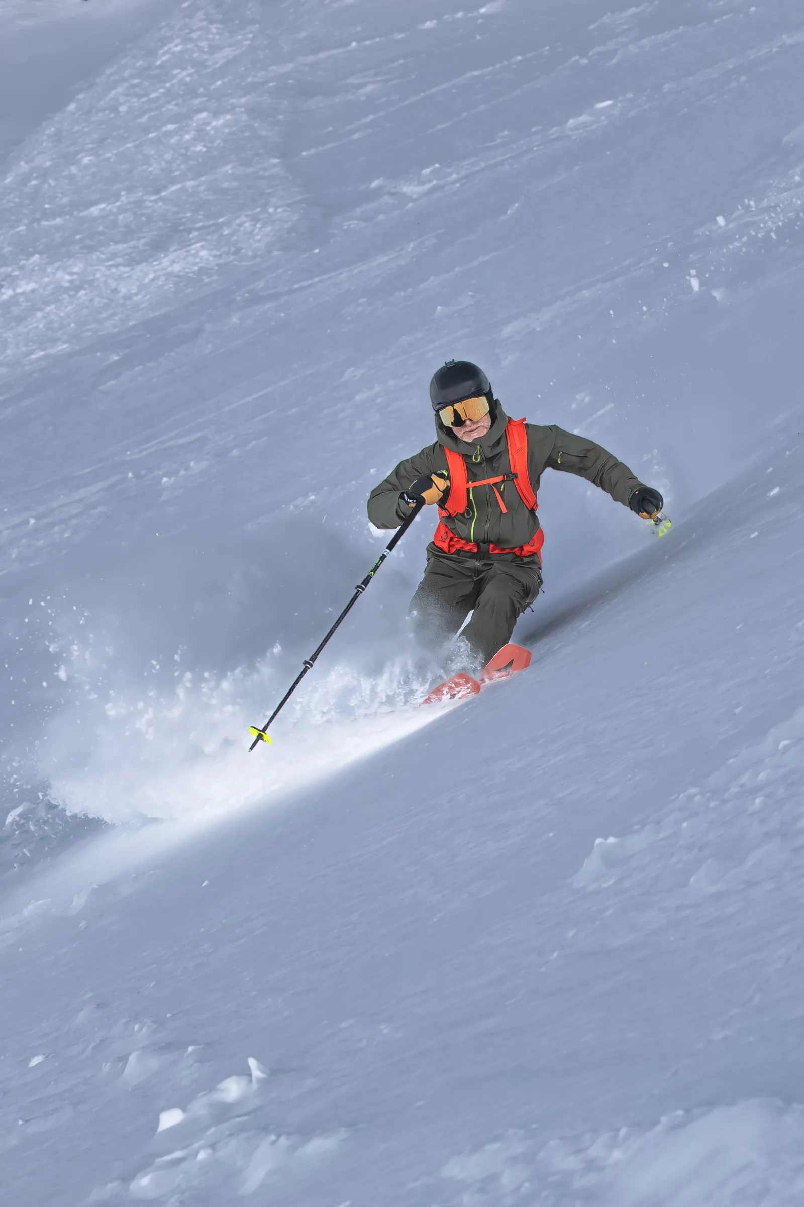 Bernd Hassmann skis through deep snow wearing a black helmet, mirrored goggles and an orange backpack, with snow spraying around his red ski tips.