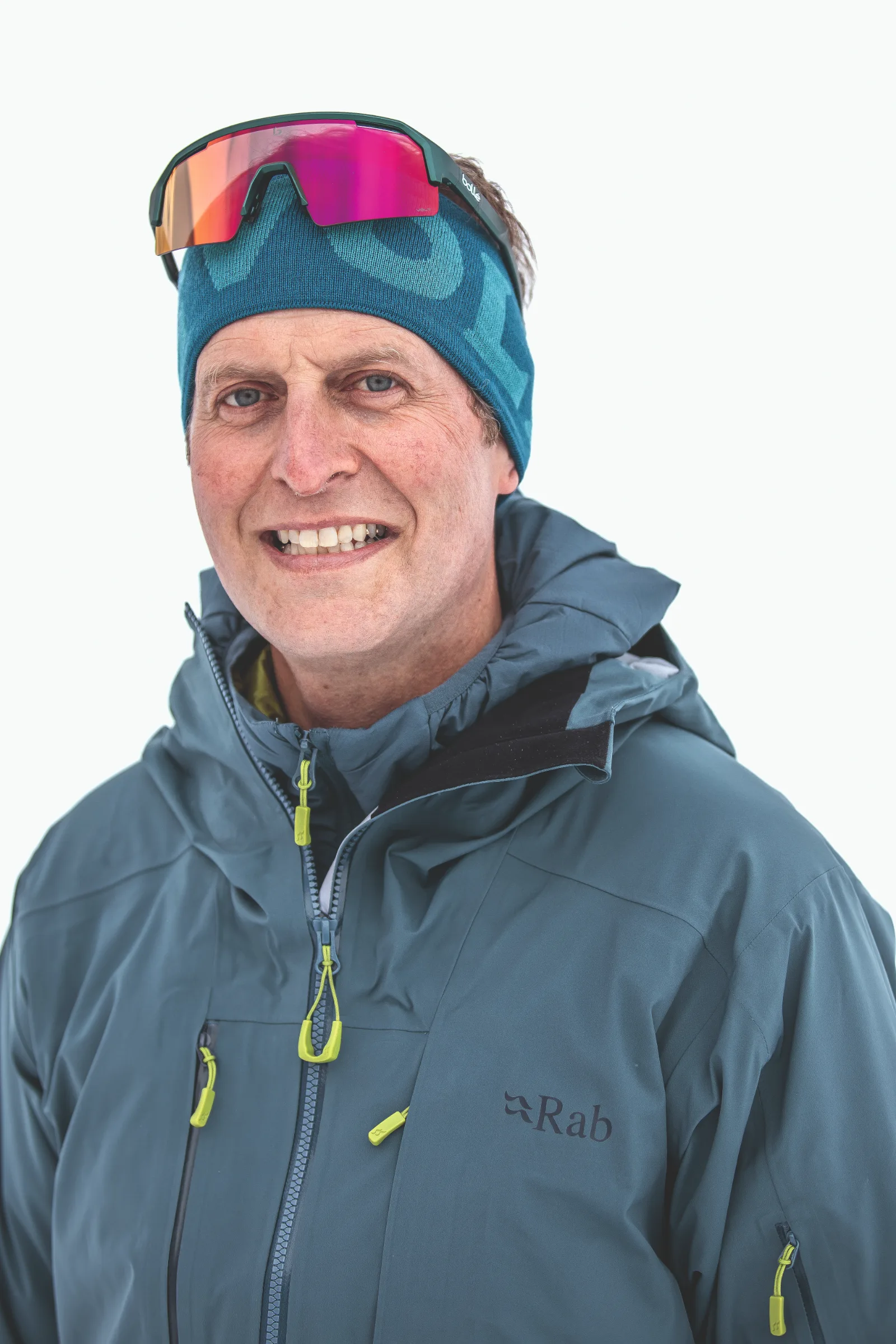 Christoph Öfele lächelt vor weißem Hintergrund in die Kamera, mit blauem Stirnband, darauf sitzender verspiegelter Sportbrille und blaugrauer Jacke.