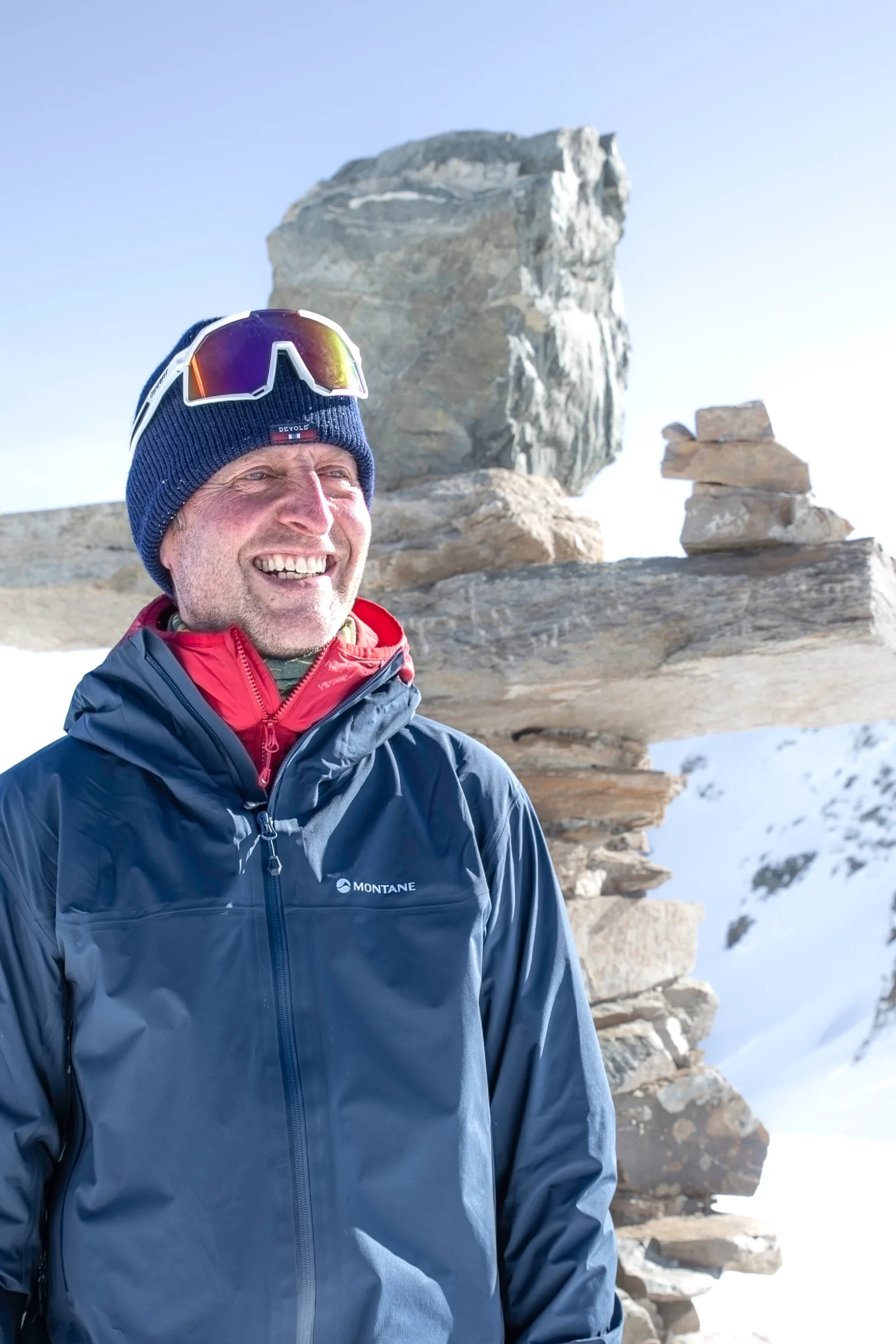 Christoph Öfele lächelt mit blauer Mütze und darauf sitzender Sportbrille vor aufgeschichteten Steinplatten und einem verschneiten Berghang.