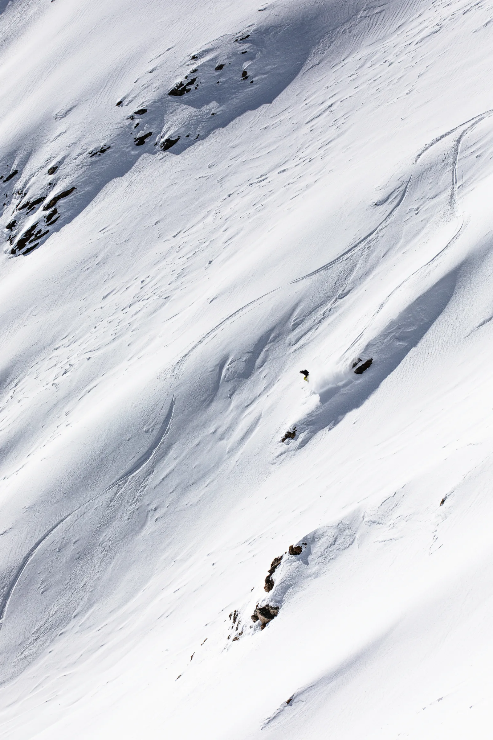 Christoph Öfele zieht auf einem weiten, steilen Schneehang bei Lenzerheide einen Schwung mit Schneefahne zwischen vereinzelten Felsen.