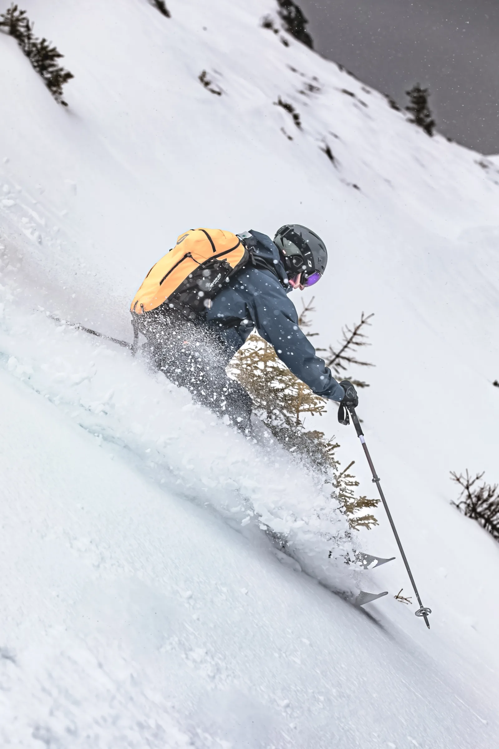 Christoph Öfele fährt mit orangefarbenem Rucksack an kleinen Nadelbäumen vorbei und wirbelt auf dem steilen Hang Schnee auf.