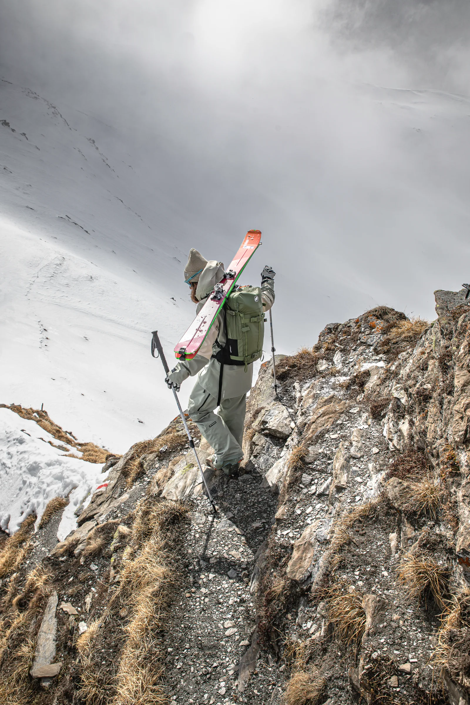 Claudia Kohler steps down a rocky path using poles, with skis strapped to her green backpack above a cloud-shrouded snowy slope.