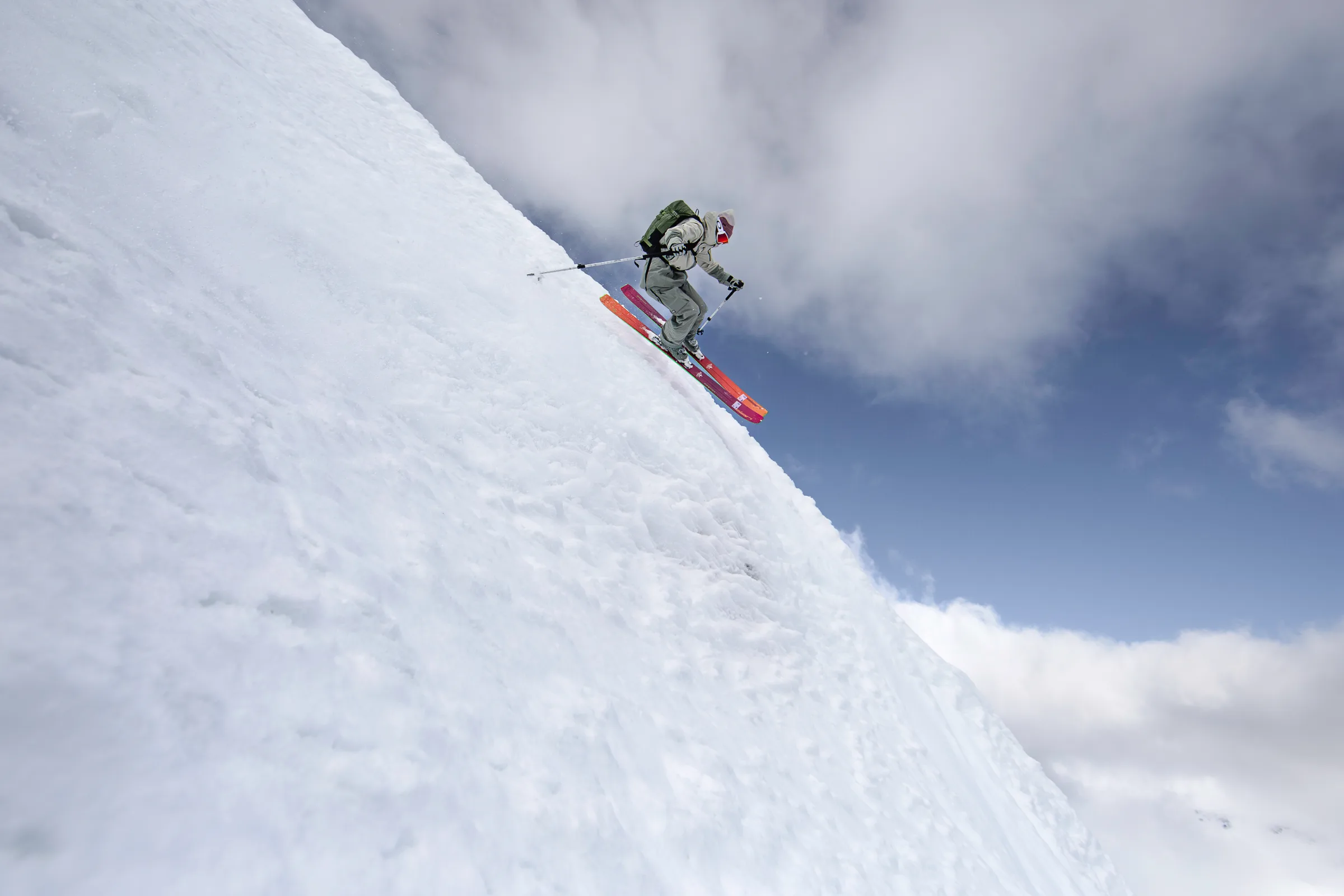 Claudia Kohler jumps with parallel skis over a steep snow lip against blue sky and white clouds.