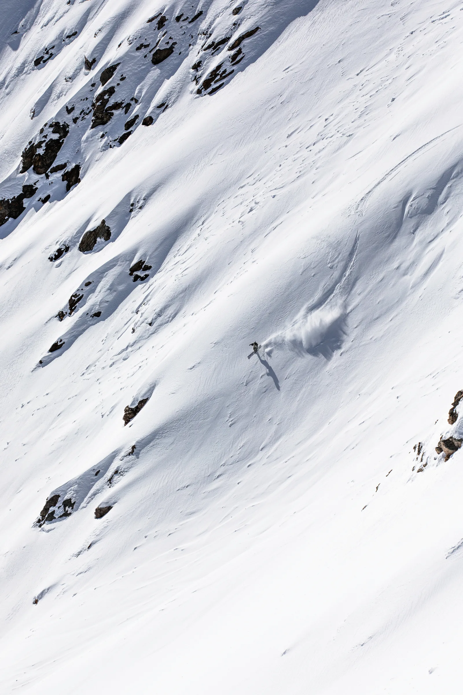 Claudia Kohler skis down a broad snowy slope between exposed rocks, trailing a large cloud of snow and casting a long shadow.
