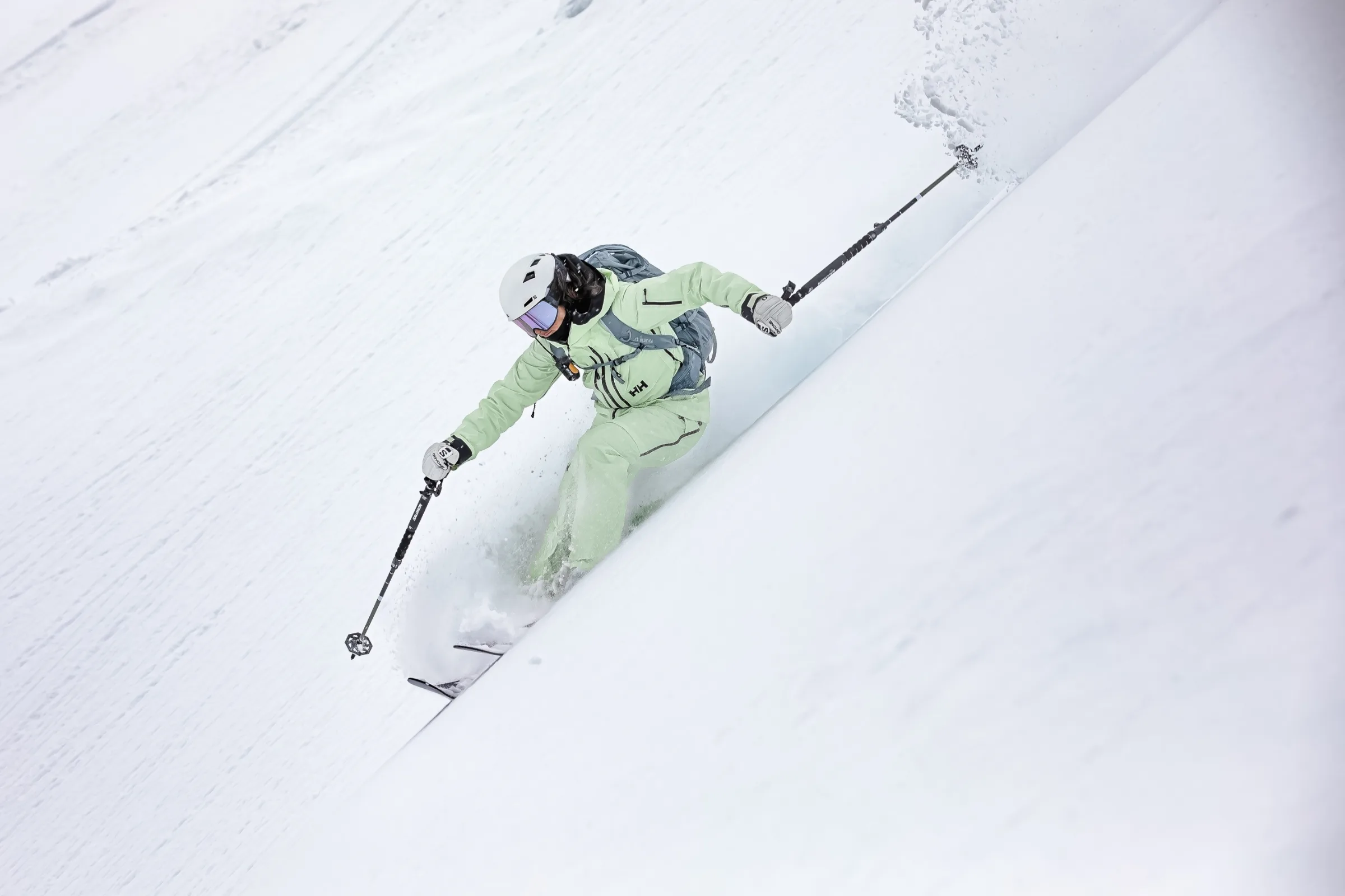 Claudia Kohler skis through deep snow on a steep slope, wearing pale green ski clothing, a white helmet and a grey backpack.