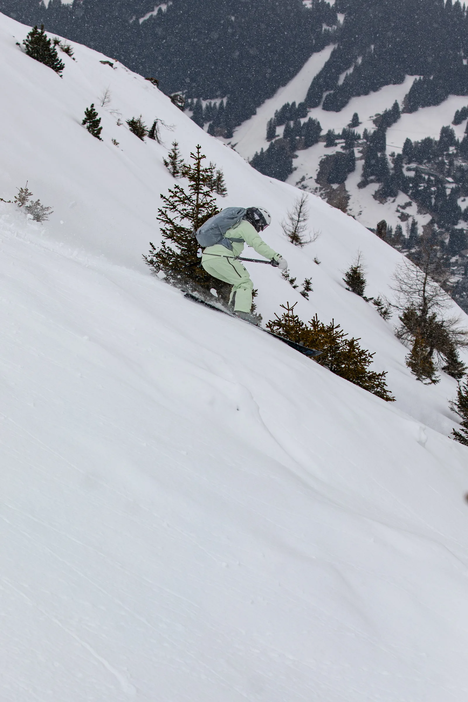Claudia Kohler skis down a steep slope between small conifers with her knees bent as snow falls.