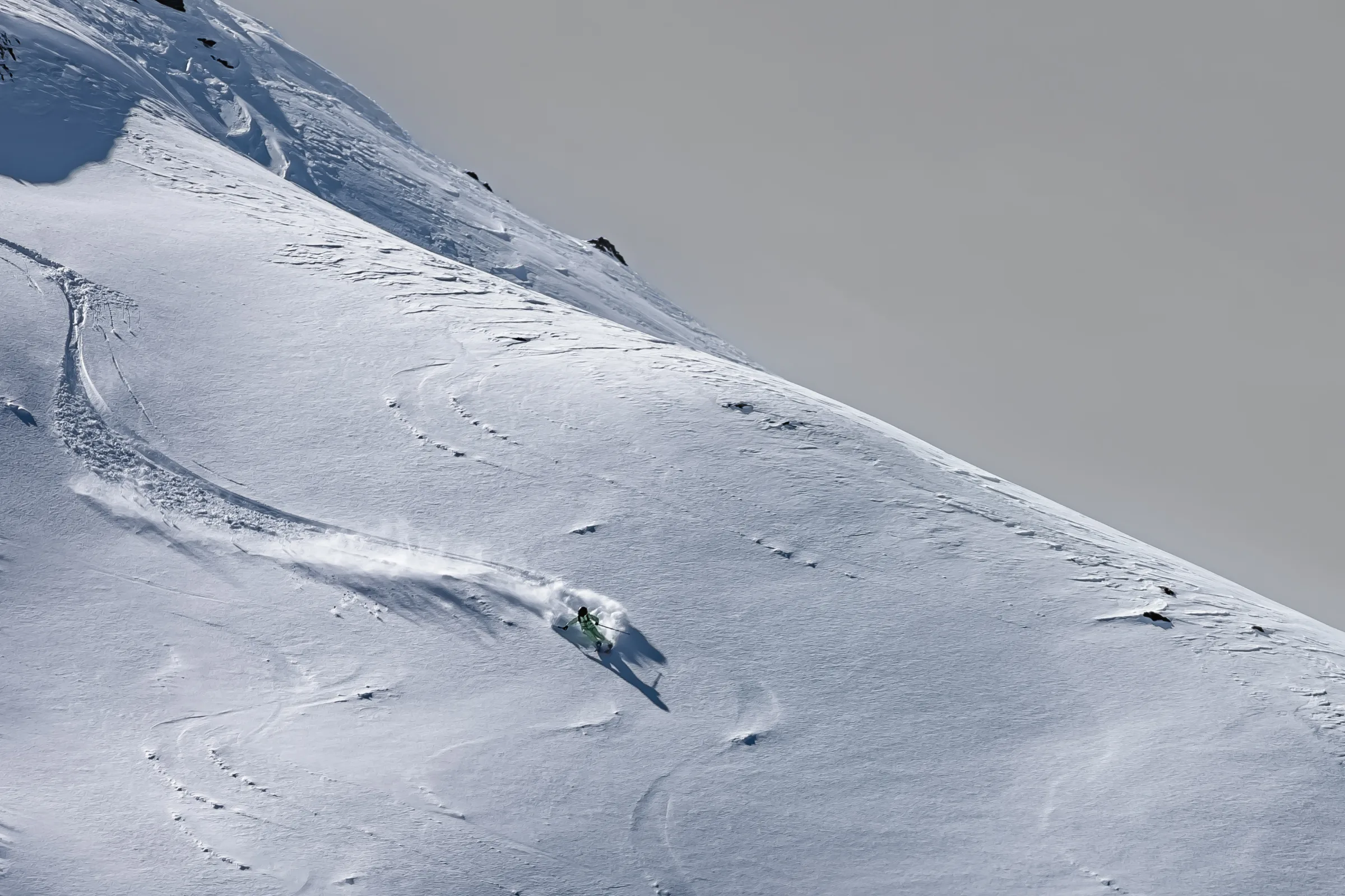 Claudia Kohler leans into a turn on an open snowy slope, with snow spraying behind her and a long shadow stretching downhill.