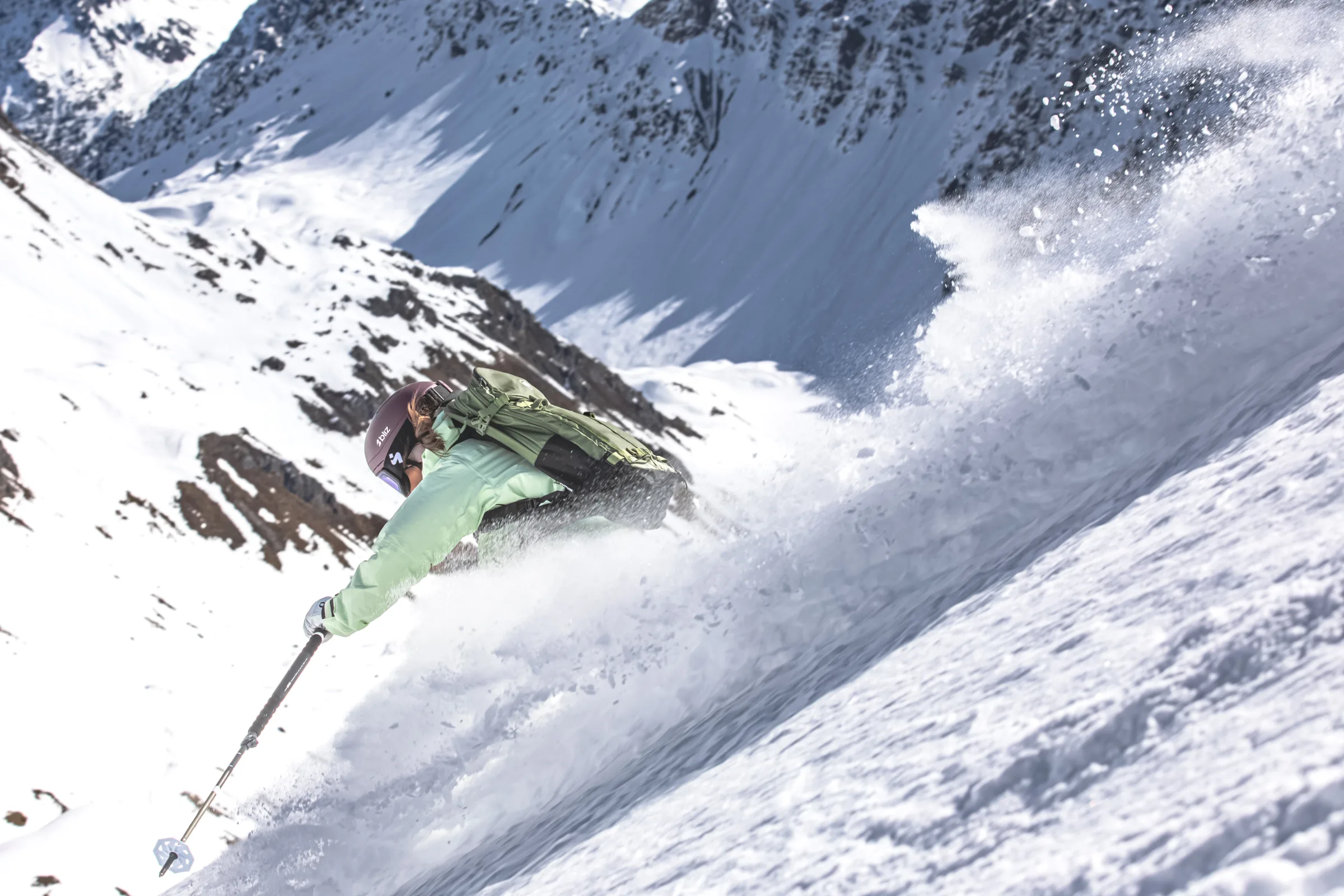 Seen from behind, Claudia Kohler leans low into a turn with a green backpack as snow sprays high against a mountain backdrop.