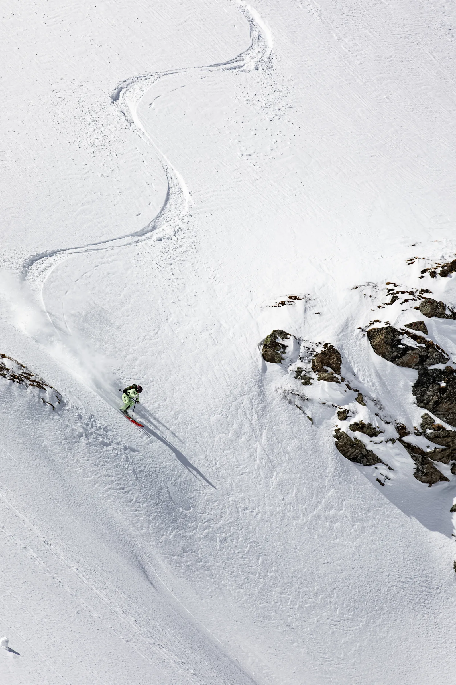 Claudia Kohler skis down a sunlit snowy slope beside rocks, with her ski track winding through several turns above her.