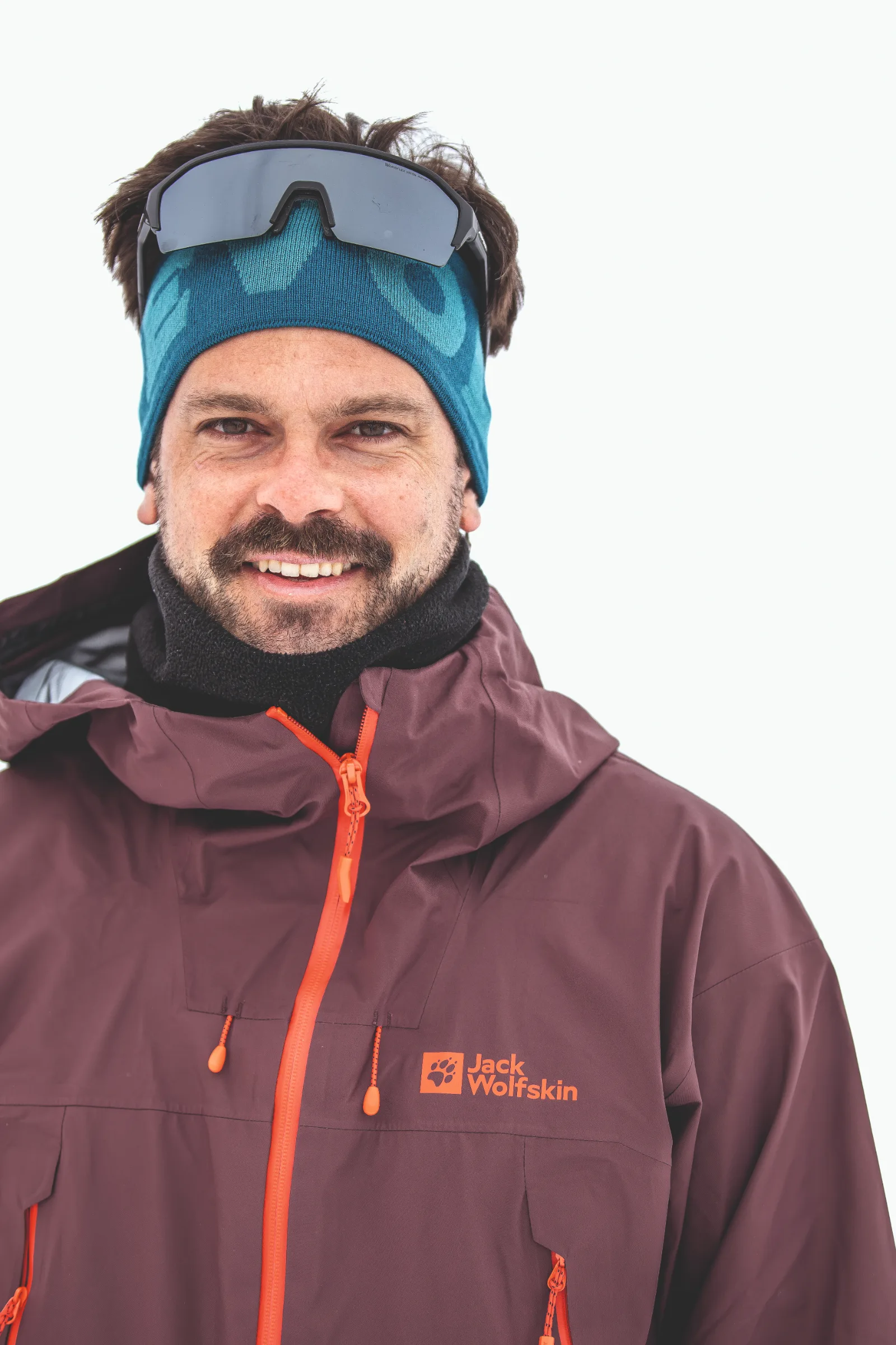 Florian Eigler smiles at the camera against a white background, wearing a blue headband with sunglasses perched on top and a burgundy jacket with an orange zipper.