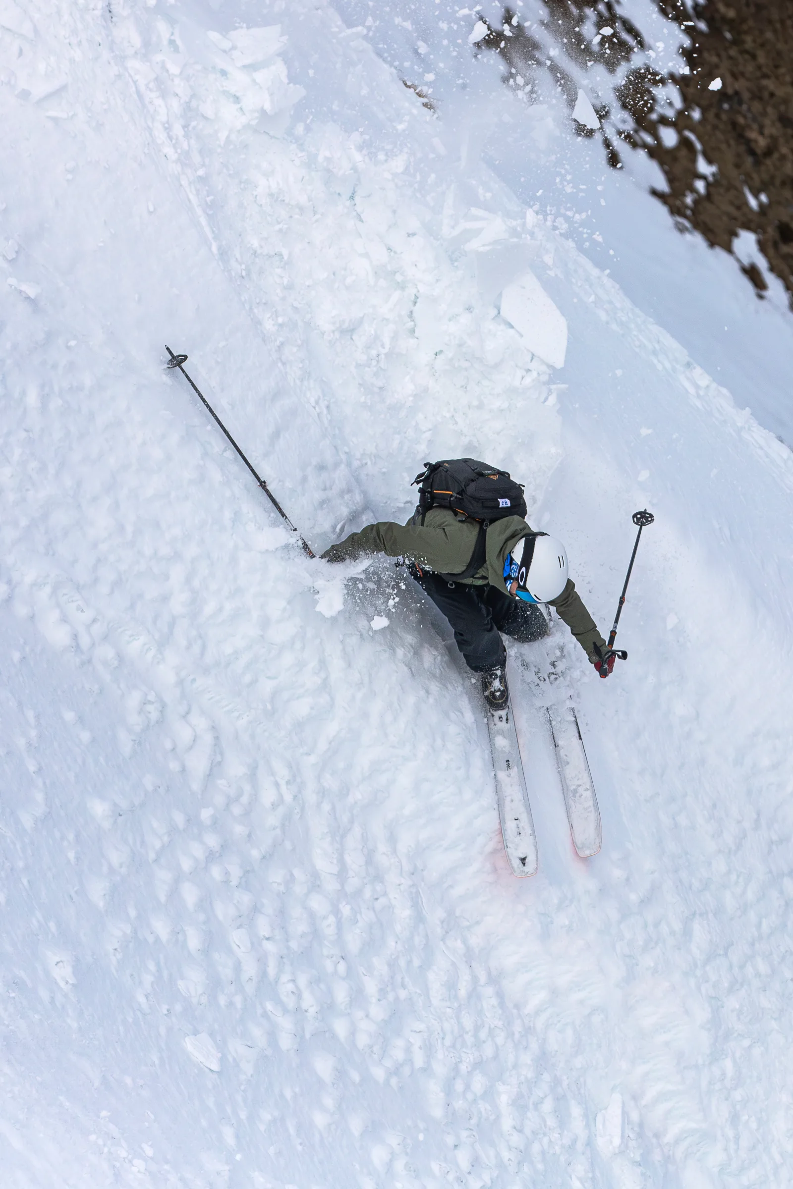 Seen from above, Florian Eigler skis in a deep crouch through flying snow beside a rock wall, wearing a white helmet and backpack.