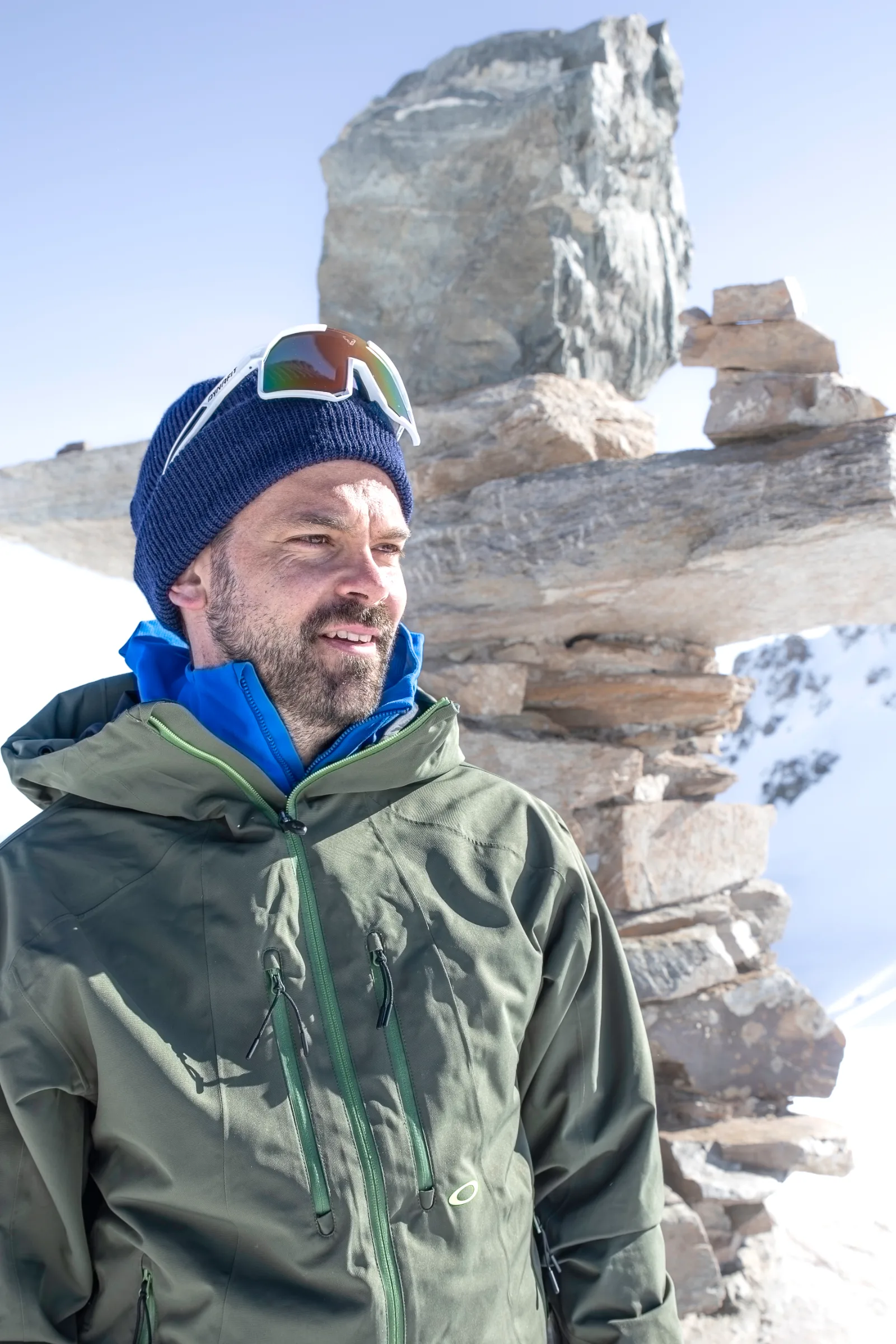 Florian Eigler smiles and looks aside, wearing a blue beanie with sunglasses perched on top, with stacked stones in the snow behind him.
