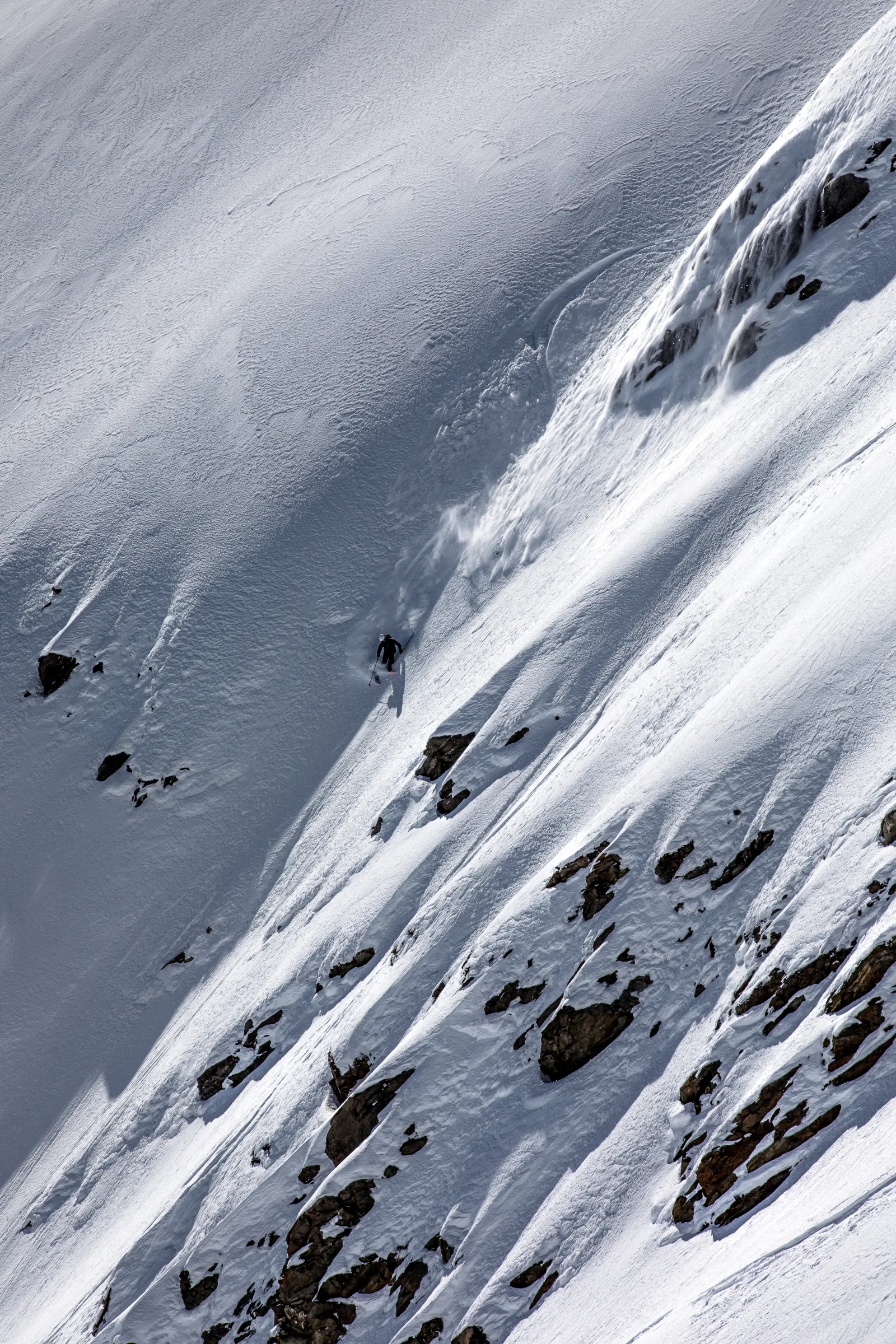 Florian Eigler descends a steep snowy slope between exposed rocks below a curved ski track, trailing a plume of snow.