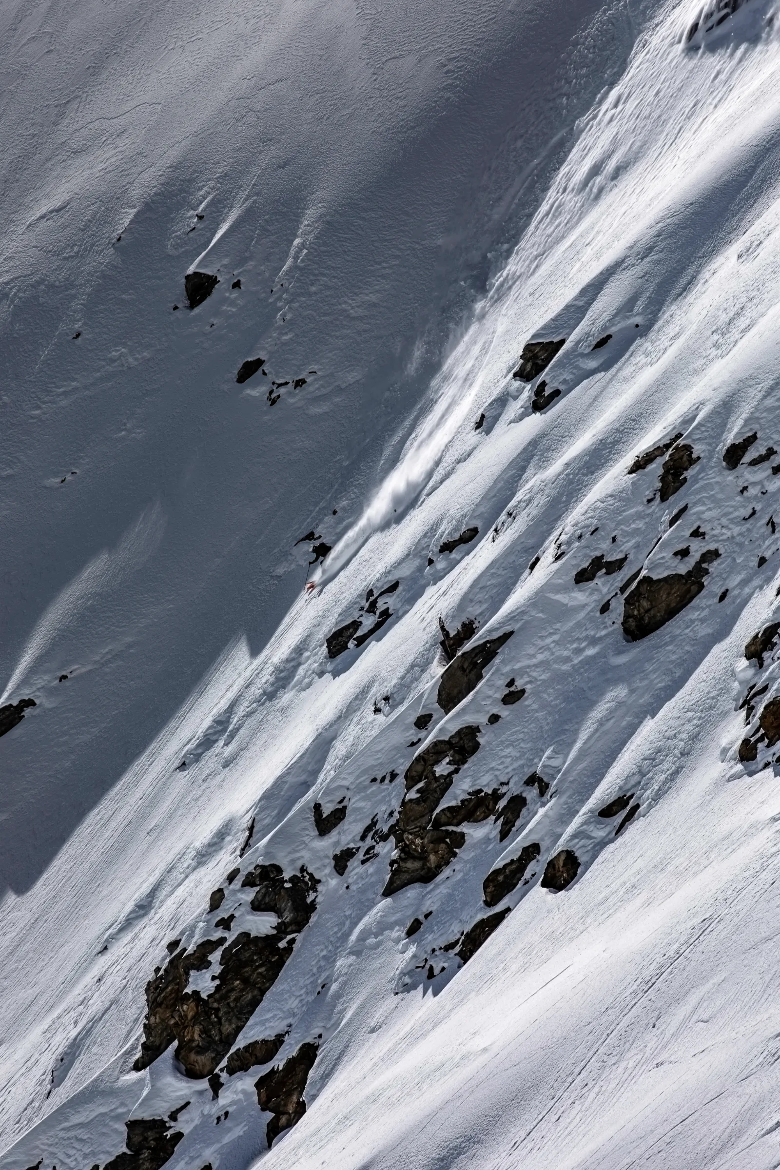 Florian Eigler skis downhill beside a rock-studded section of slope, with a long plume of snow trailing up the steep face.
