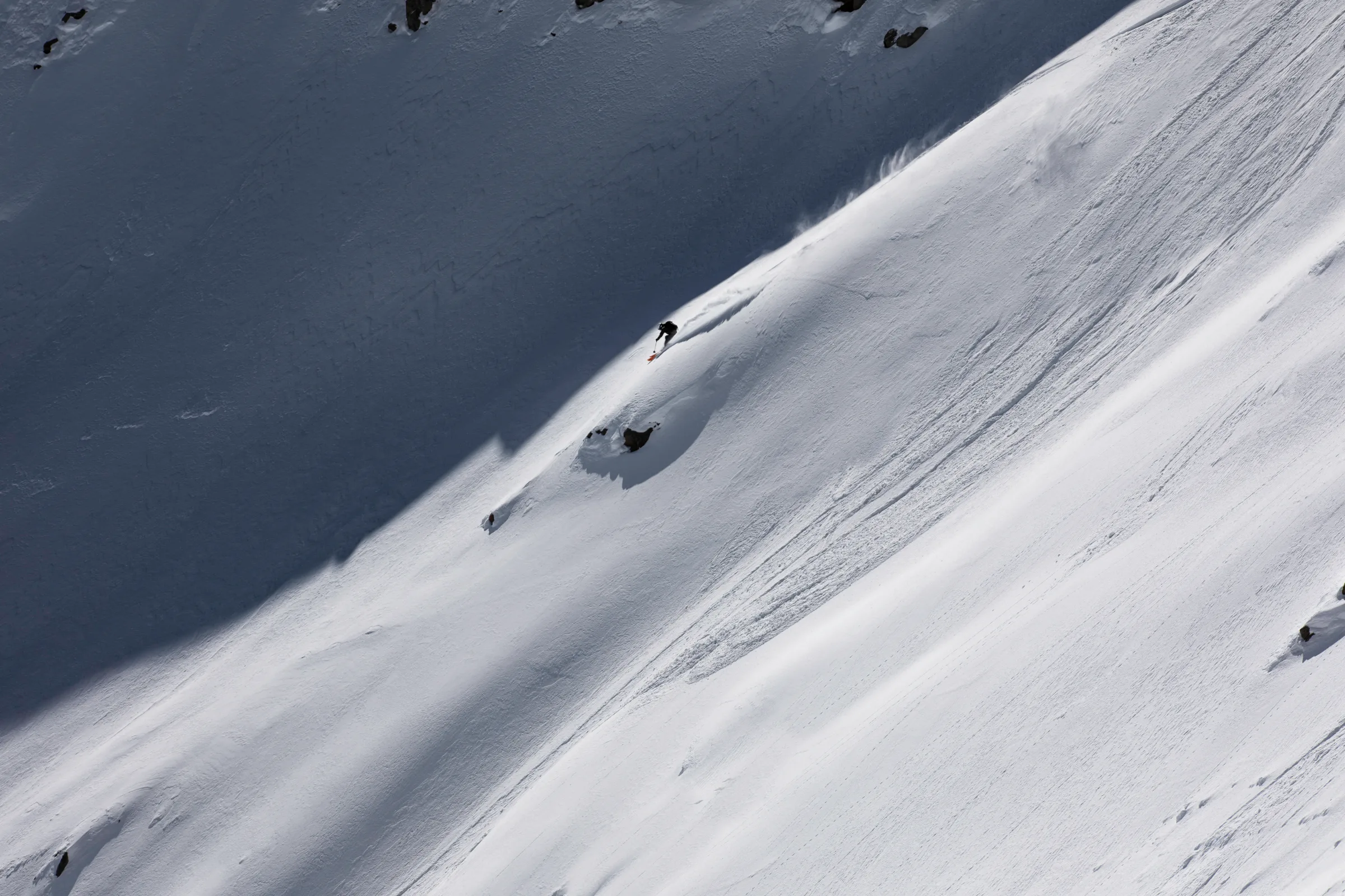 Florian Eigler skis across a broad snow slope along the boundary of sunlight and shade, leaving a track above scattered rocks.