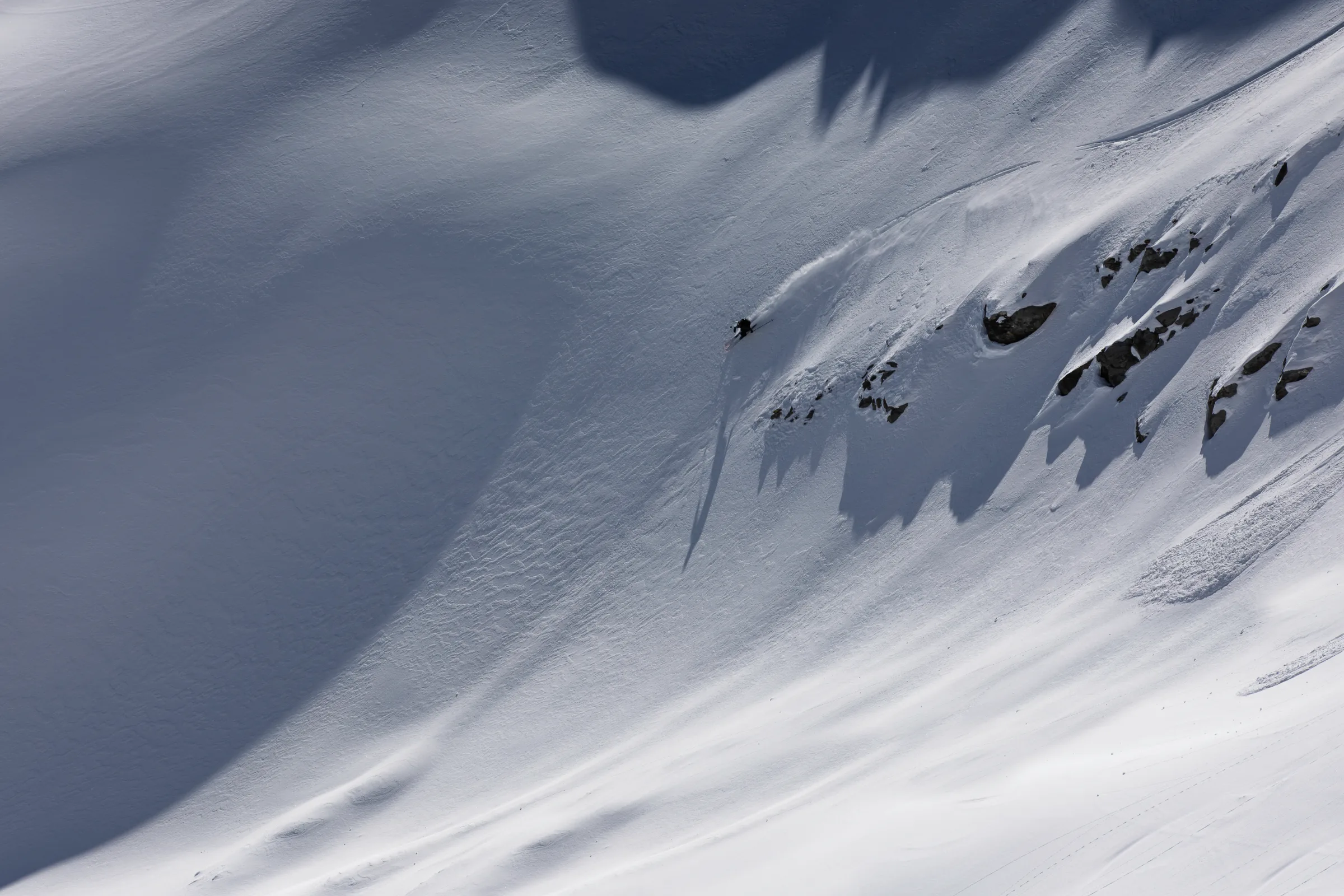 Florian Eigler turns to the left of a cluster of rocks casting long shadows across the snowy slope, spraying snow behind him.
