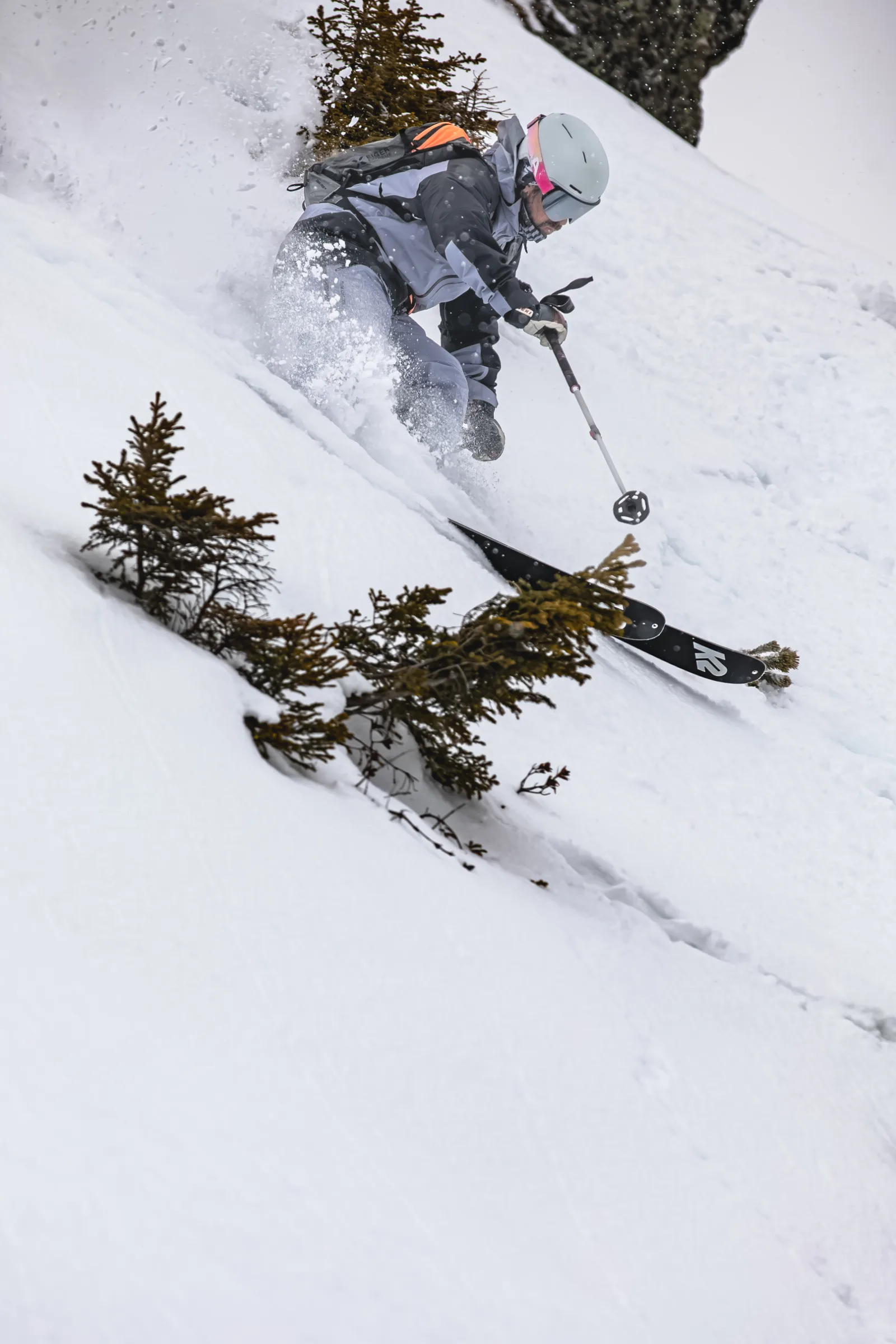 Florian Eigler crouches low as he skis down a steep snowy slope between small conifers, throwing up snow.