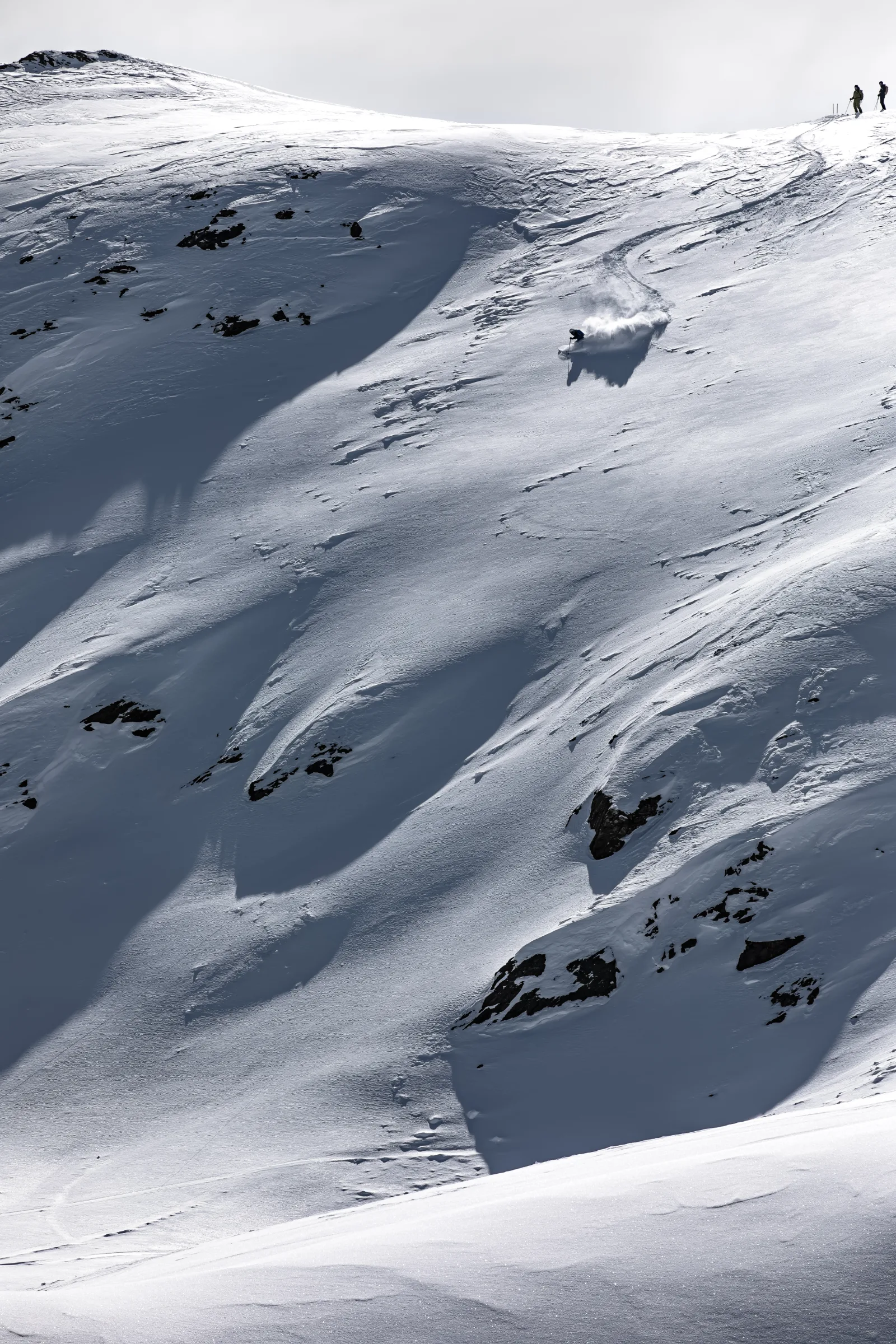 Florian Eigler makes a turn with a plume of snow on a broad, rocky slope in Lenzerheide while two people stand on the ridge above.