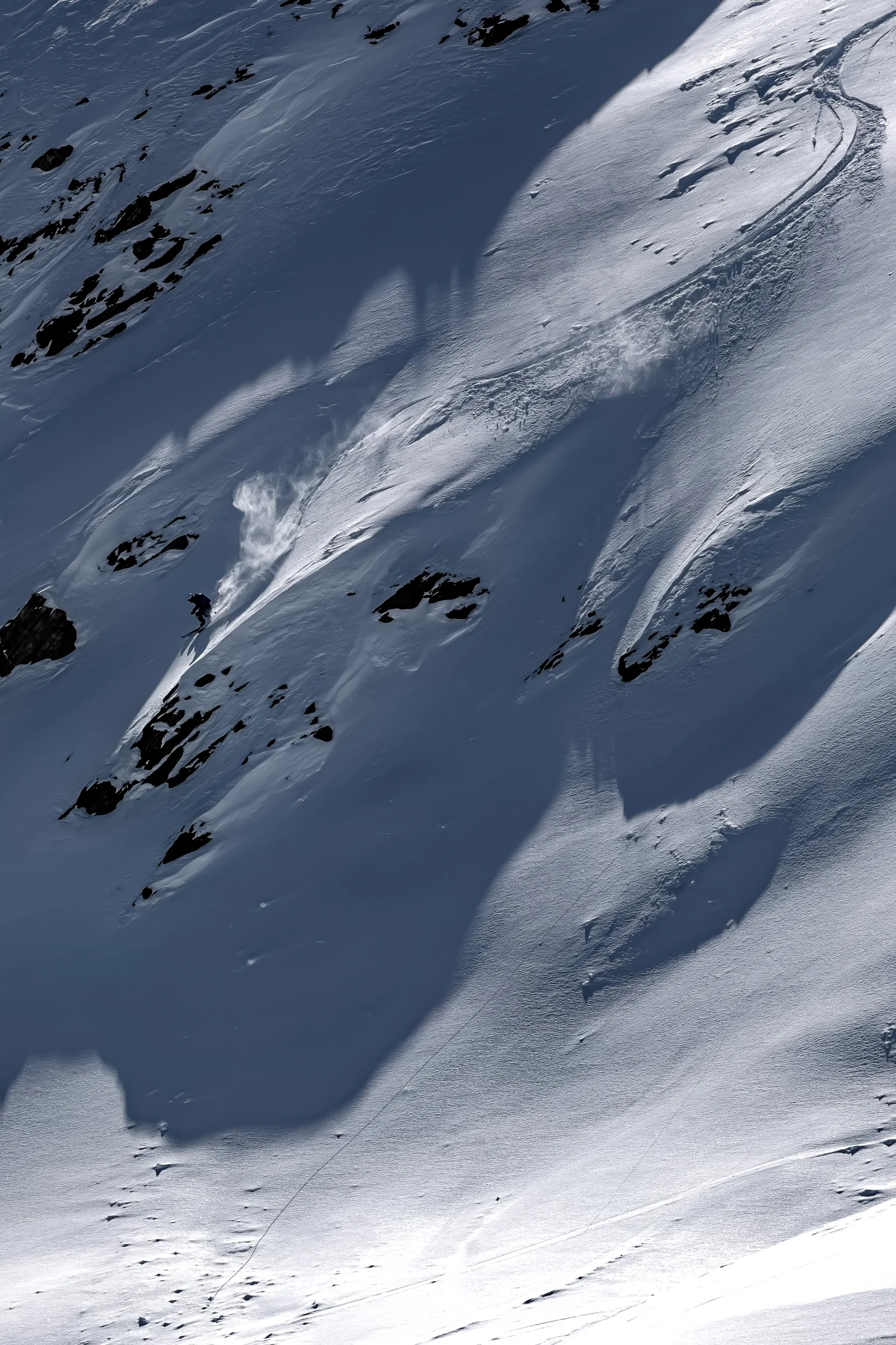 Florian Eigler skis between exposed rocks on a steep, partly shaded snow slope, with a long curved track and drifting spray behind him.