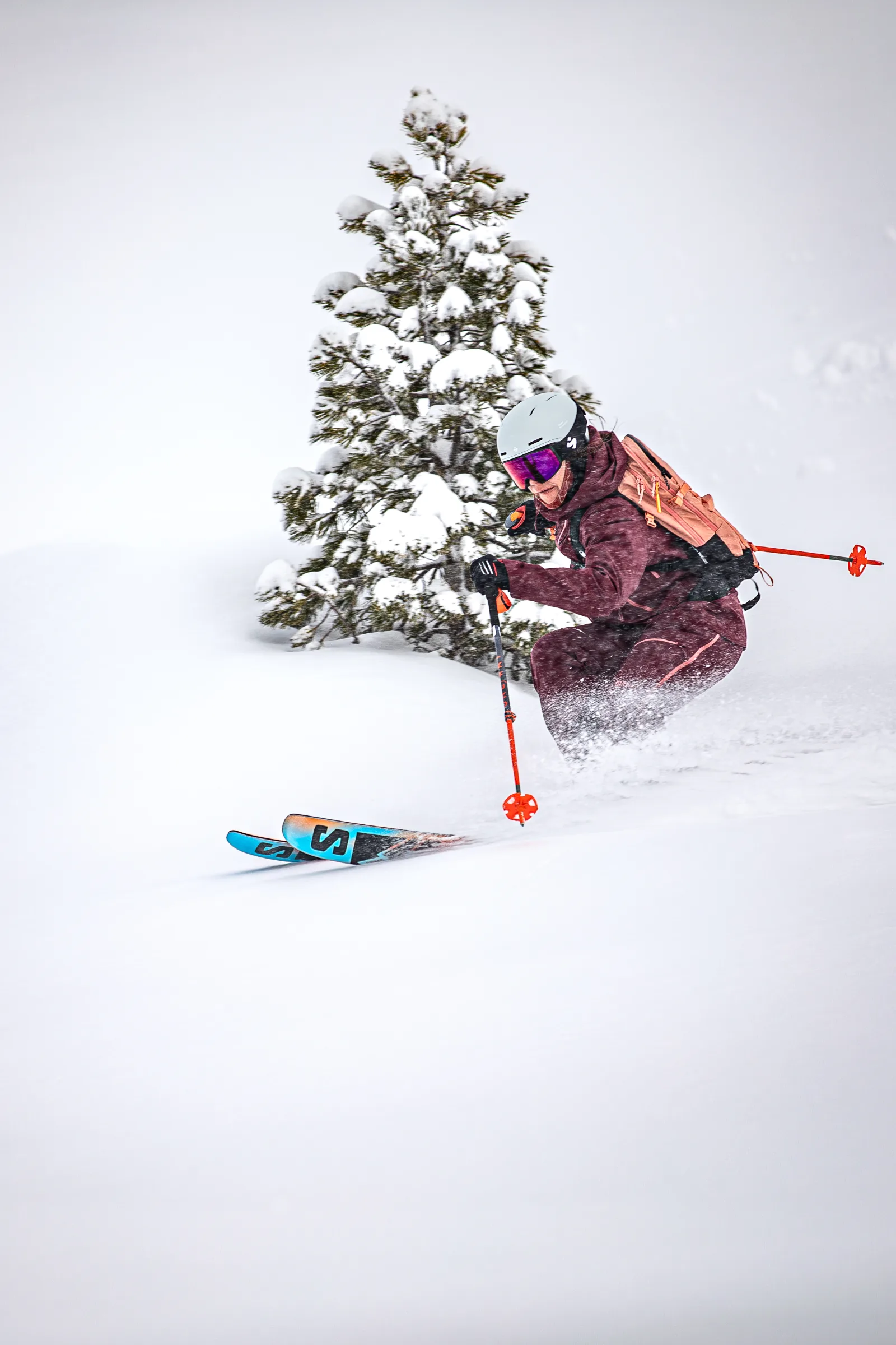 Martina Wengenmeir fährt mit Helm und korallfarbenem Rucksack durch tiefen Schnee an einem verschneiten Nadelbaum vorbei, während Schnee um ihre Knie spritzt.