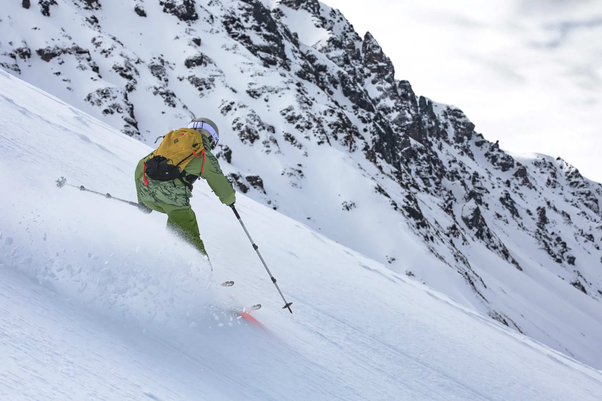 Martina Wengenmeir fährt in grüner Skikleidung mit gelbem Rucksack einen steilen Schneehang vor schroffen Felswänden in Lenzerheide hinab.